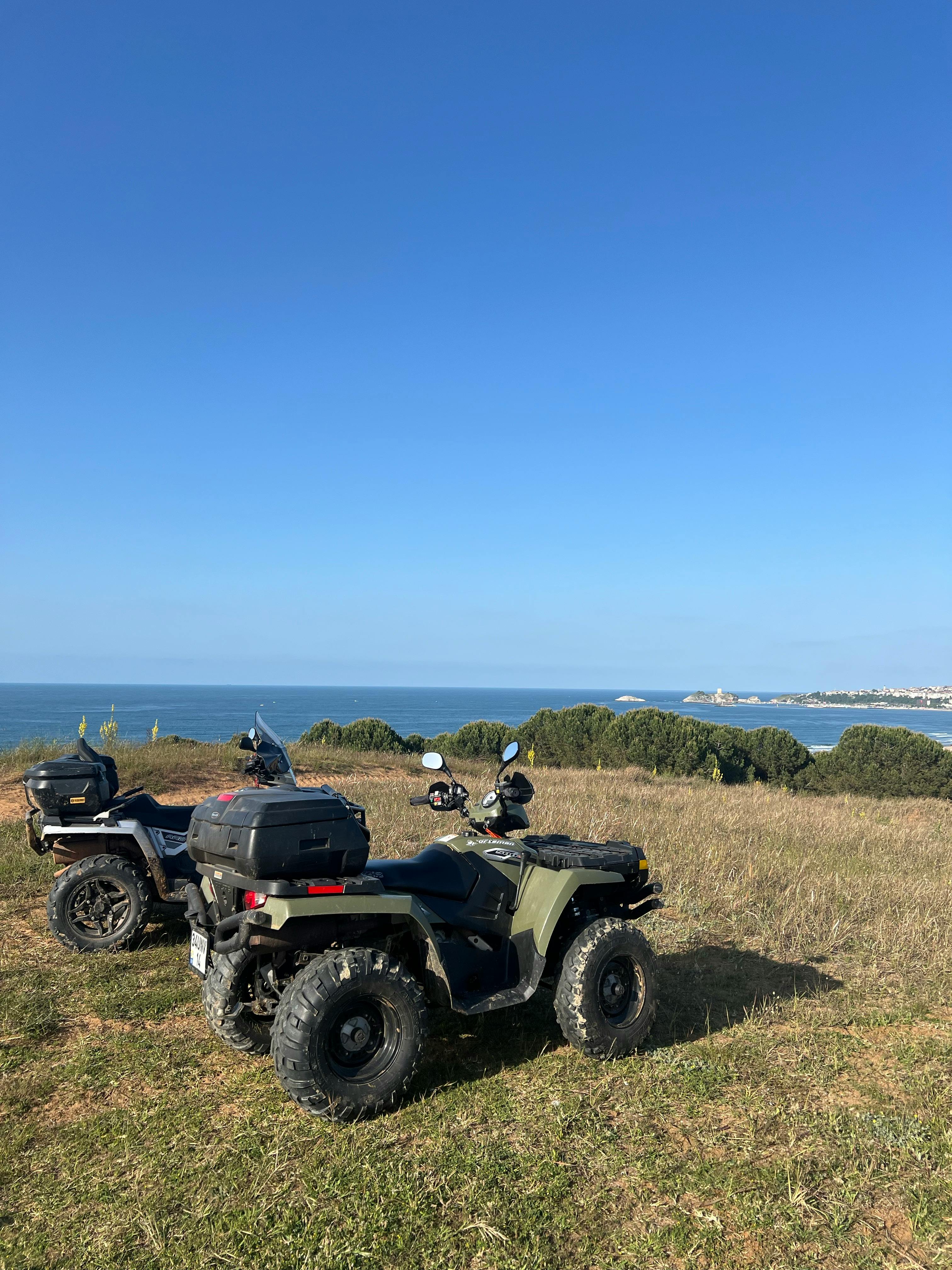 Two ATVs parked on a coastal hillside with a beautiful ocean view under clear blue skies.
