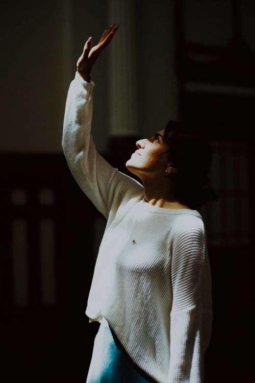 Free A woman in a white sweater reaches up towards a beam of light indoors, embodying contemplation and aspiration. Stock Photo