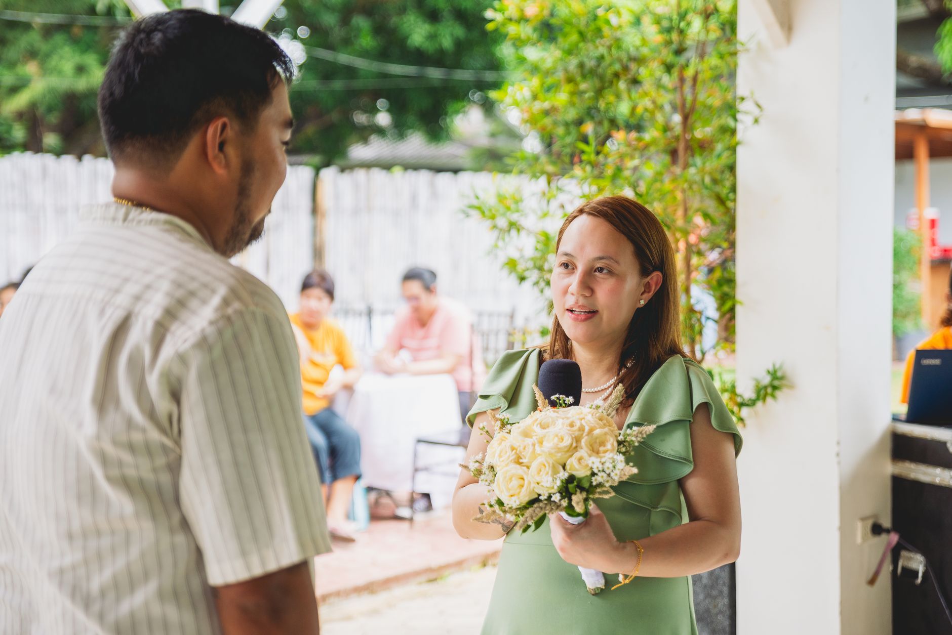 A woman gives a speech during an outdoor wedding ceremony, holding bouquets.