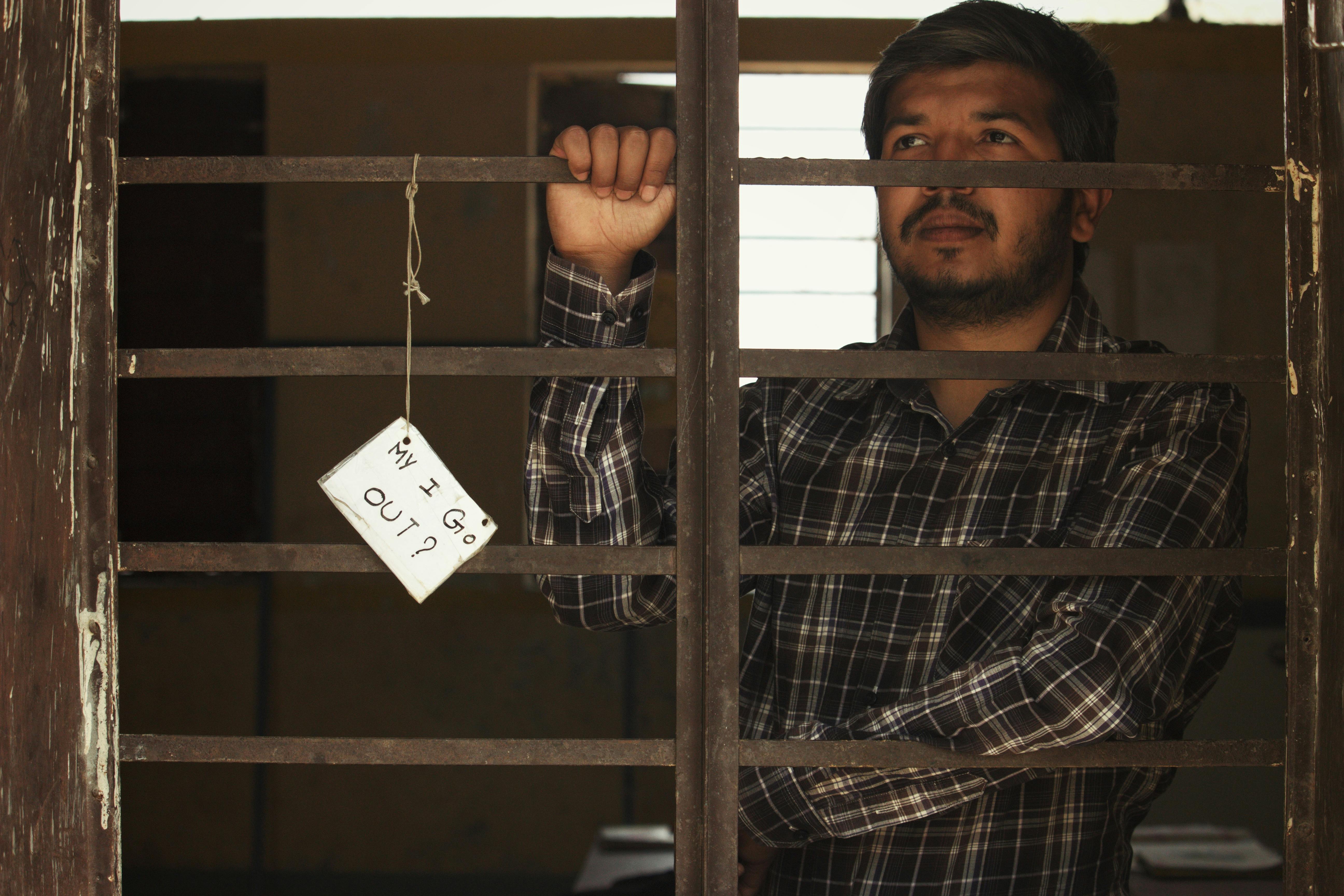 Man standing behind bars holding a sign asking 'May I Go Out?' Indoor setting with dramatic tone.