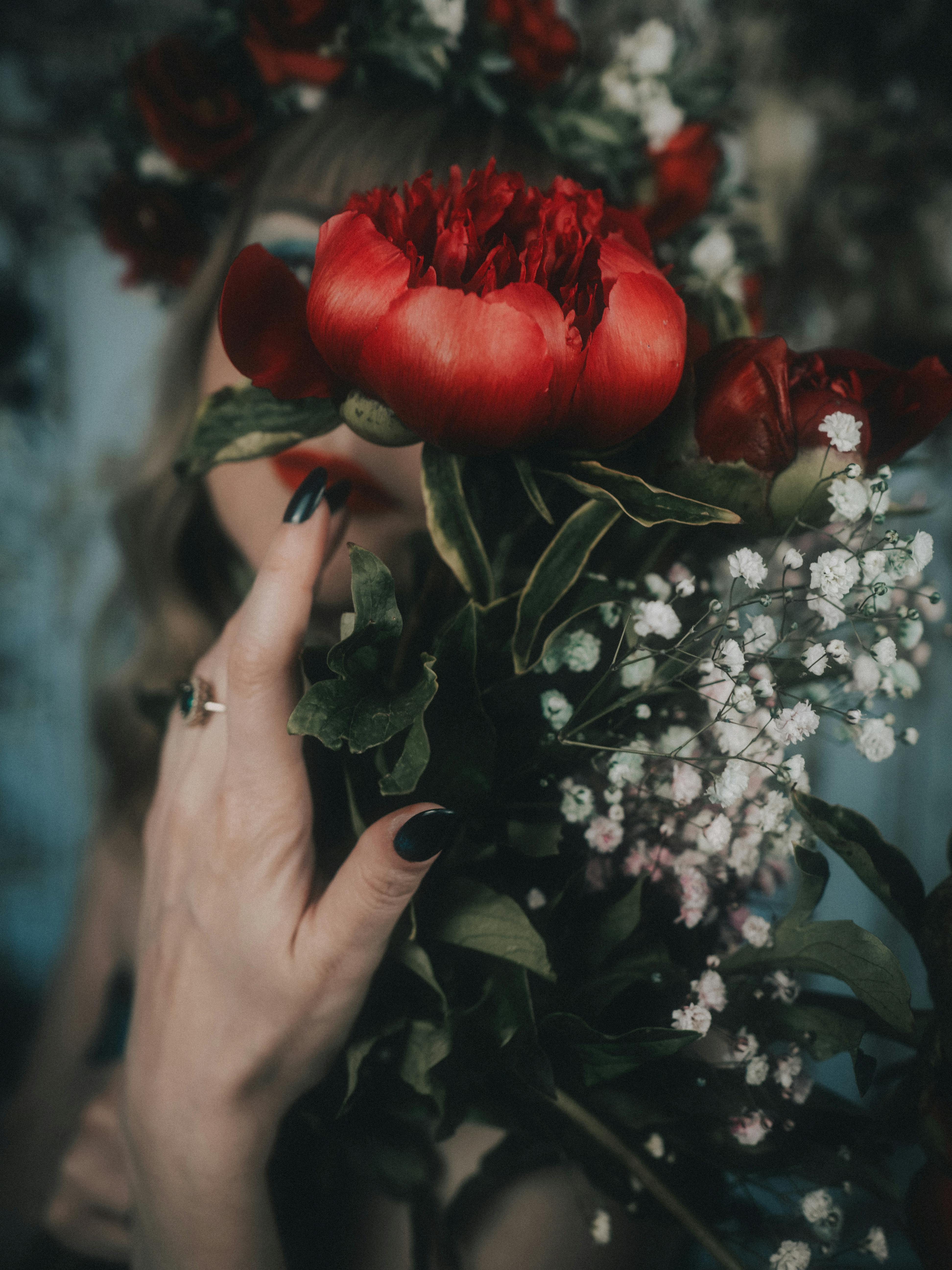A woman's hand holding a lush bouquet of red peonies and greenery, reflecting a moody and elegant vibe.