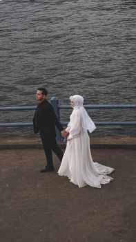 A newlywed couple holding hands and walking by the seaside, capturing a moment of love and togetherness.
