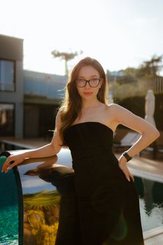 Portrait of a young woman in a black dress standing by a pool, backlit by the sunset.