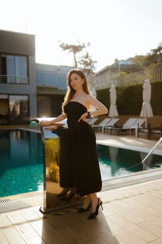 A stylish brunette woman in a black dress posing by a sunlit poolside terrace.