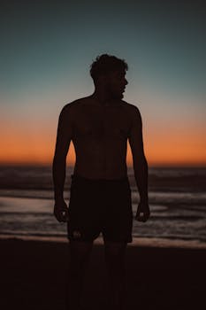 Silhouette of a shirtless man standing on a Moroccan beach at sunset, capturing a serene moment.