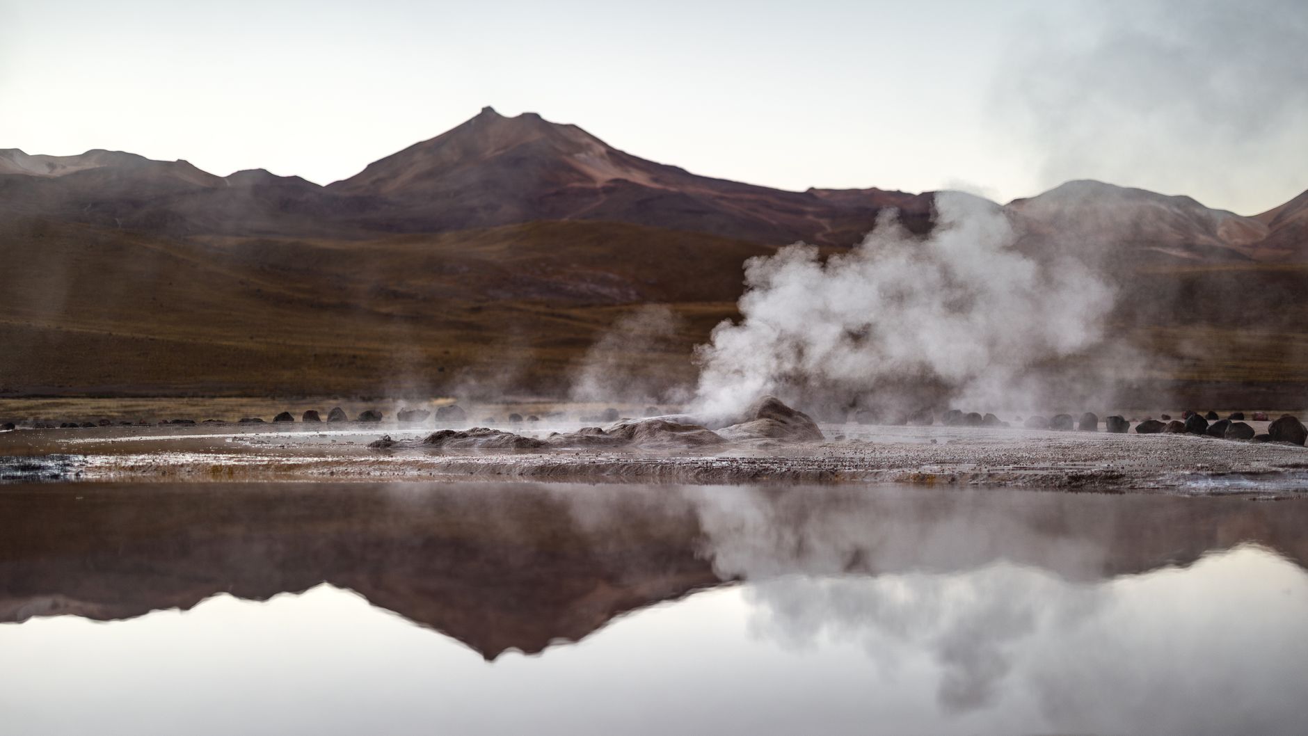 Steam columns rising from the El Tatio geyser field and reflected in a pool, with reddish Andean peaks behind