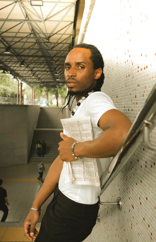 A man with long hair and a white shirt is leaning against a wall