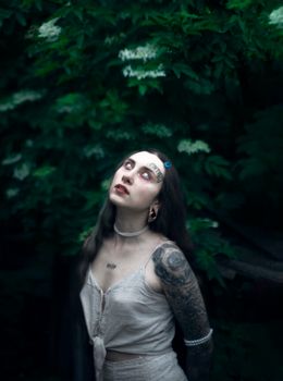 Gothic portrait of a tattooed woman amidst verdant foliage, exuding an enigmatic mood.