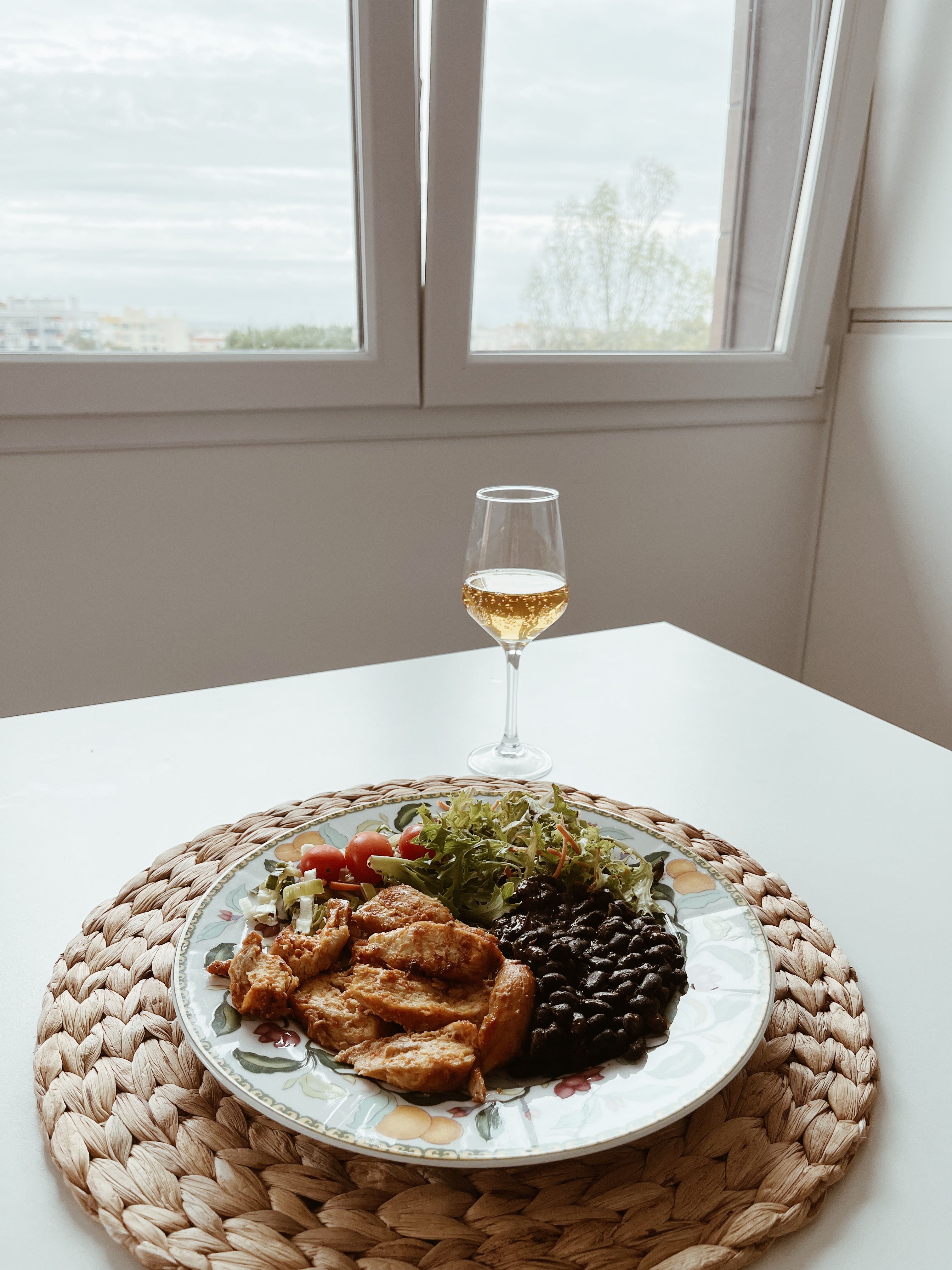 Homemade Portuguese lunch with chicken, beans, salad, and a glass of white wine by a window.