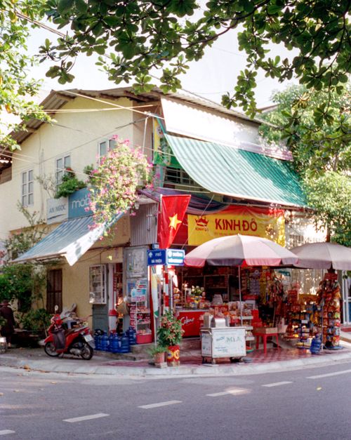 Free Vibrant street corner with traditional Vietnamese shop and red flags under lush foliage. Stock Photo