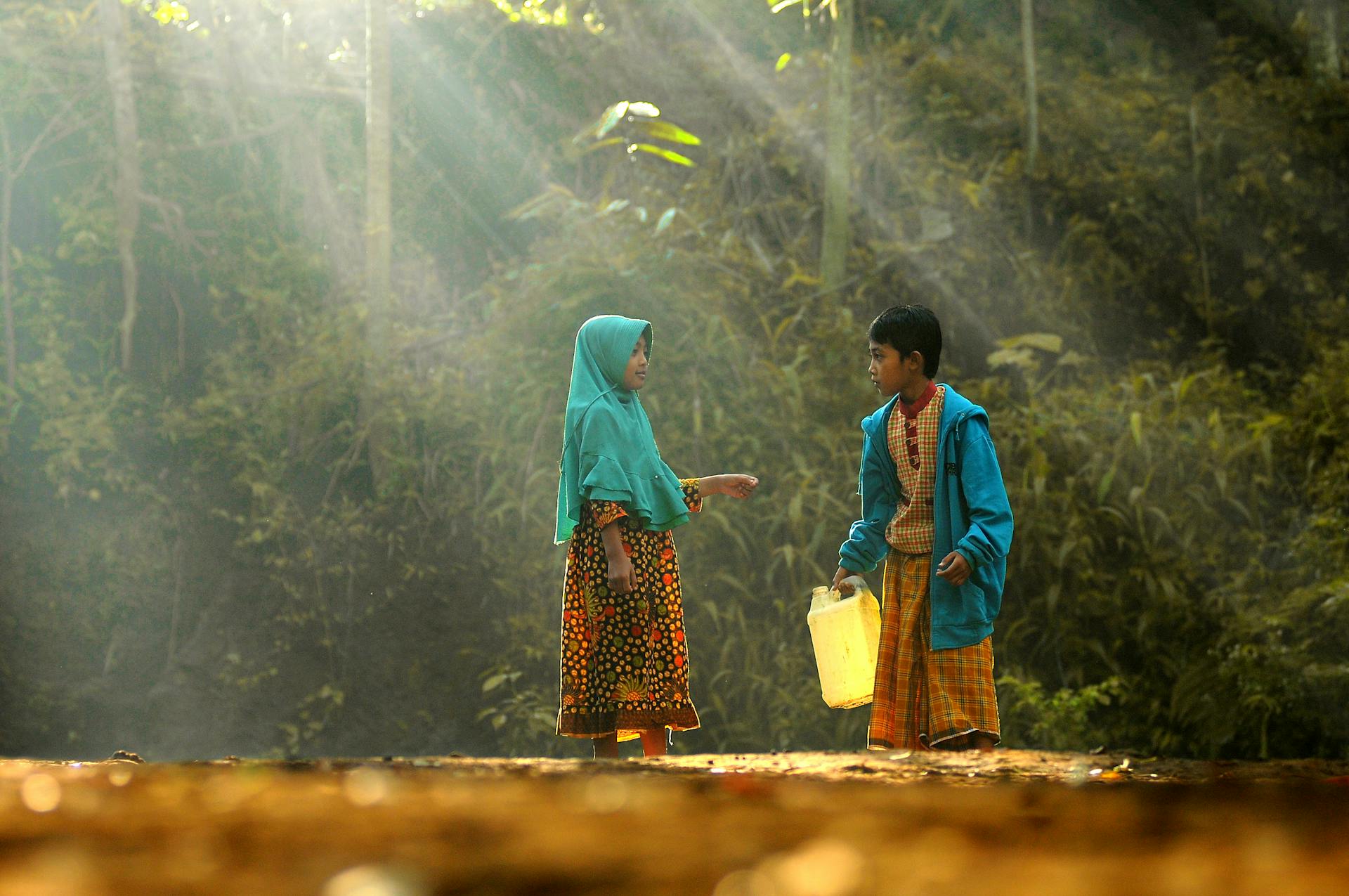 Two children stand outdoors with a jerrycan under the sunlight streaming through trees.