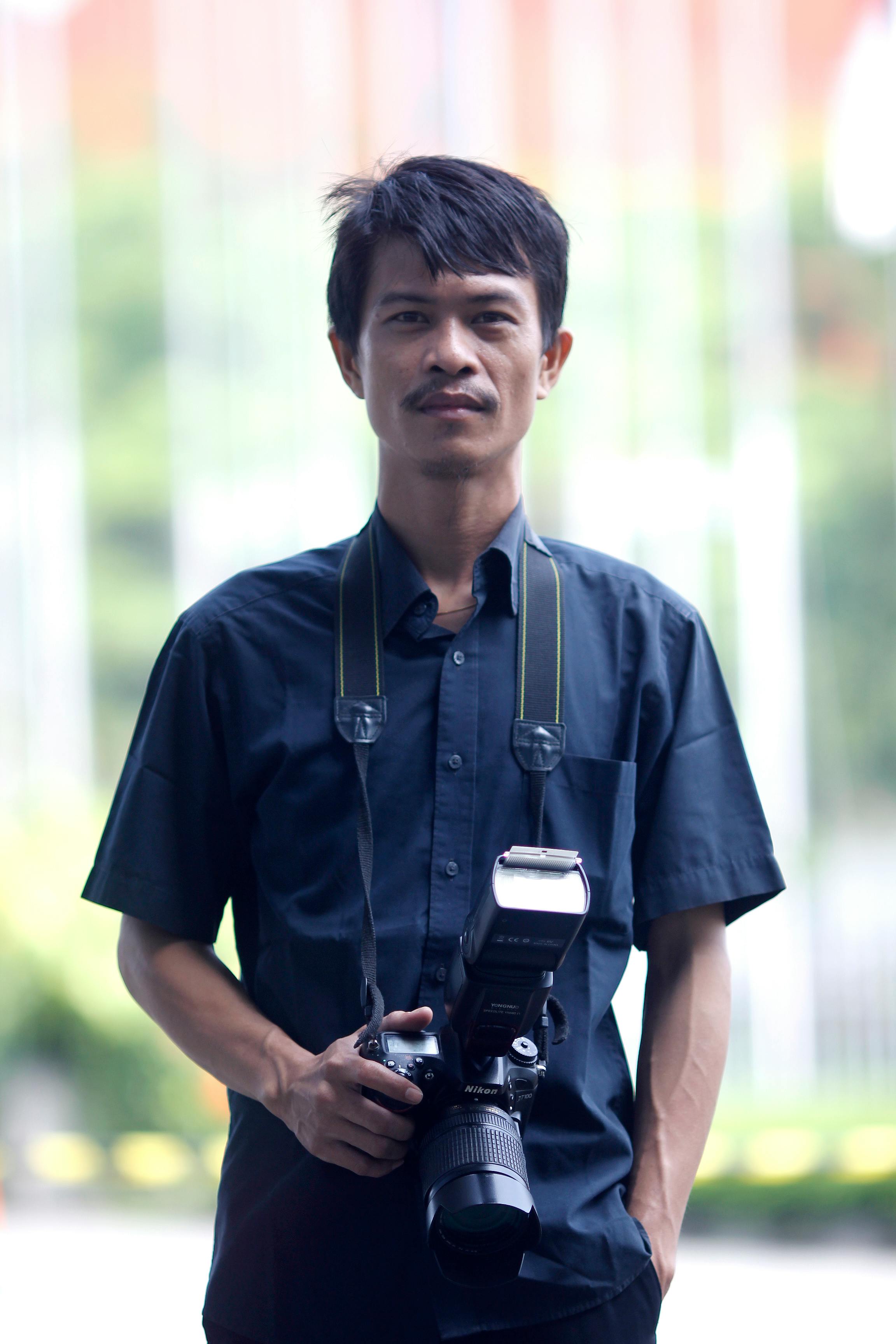 Asian male photographer in black shirt standing outdoors with camera.