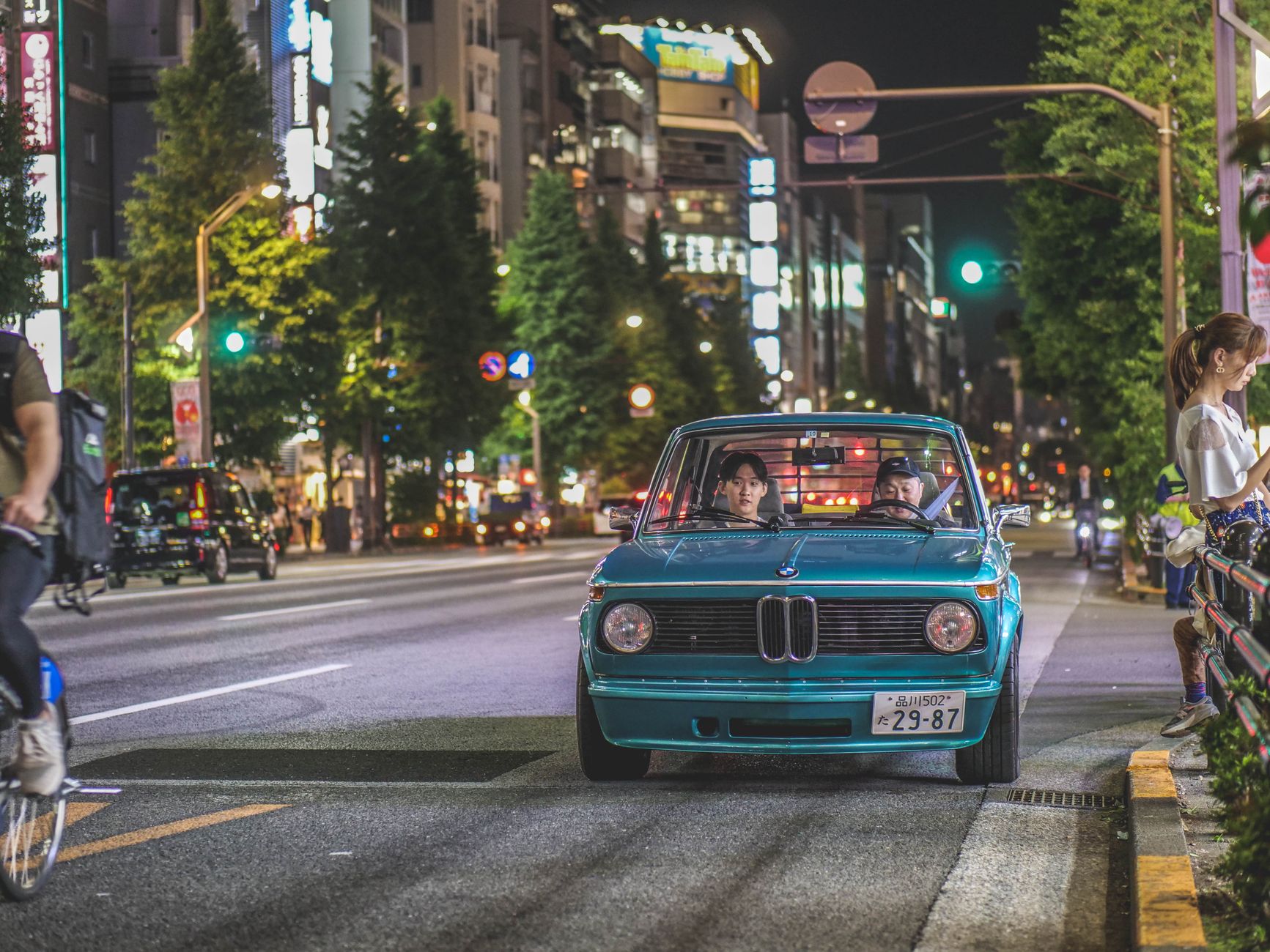 Classic BMW 2002tii driving through a vibrant city street at night.