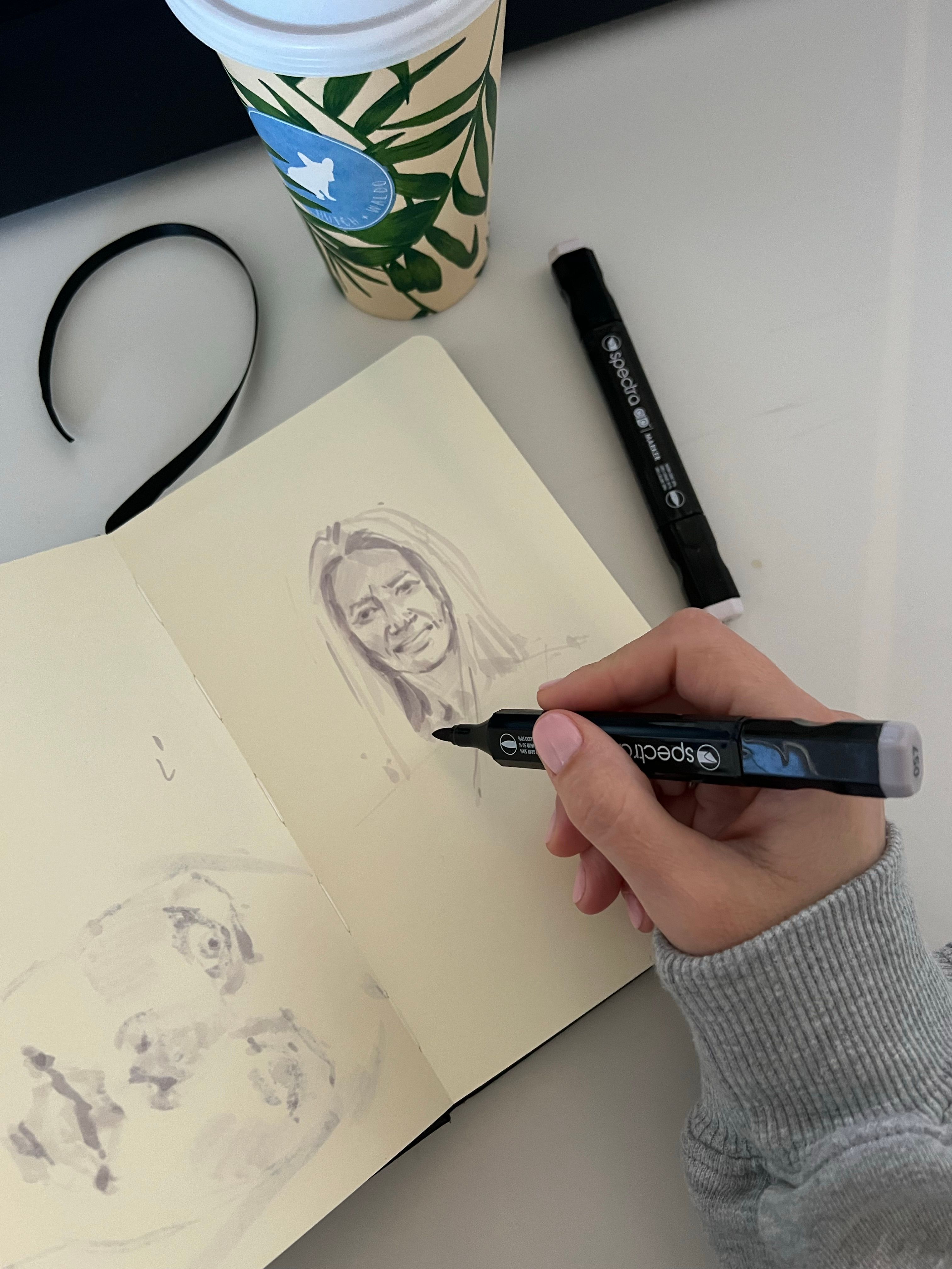 A hand drawing a portrait in a sketchbook with markers on a table.