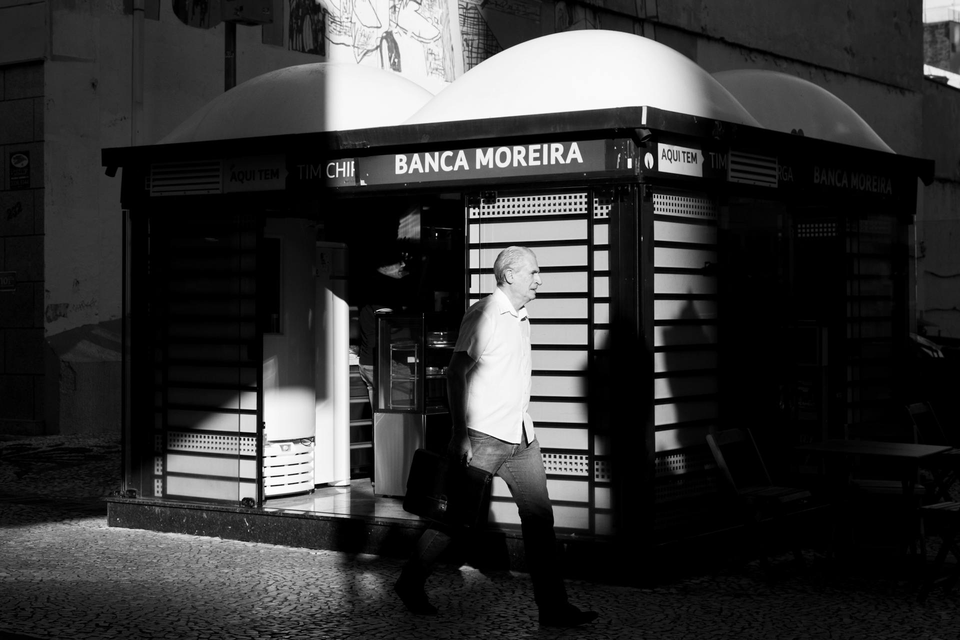 Black and white street photo of an elderly man walking past Banca Moreira kiosk in Curitiba, Brazil.