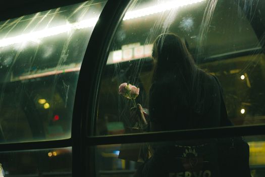 A woman with flowers waits inside a glass-covered station at night in urban Curitiba.