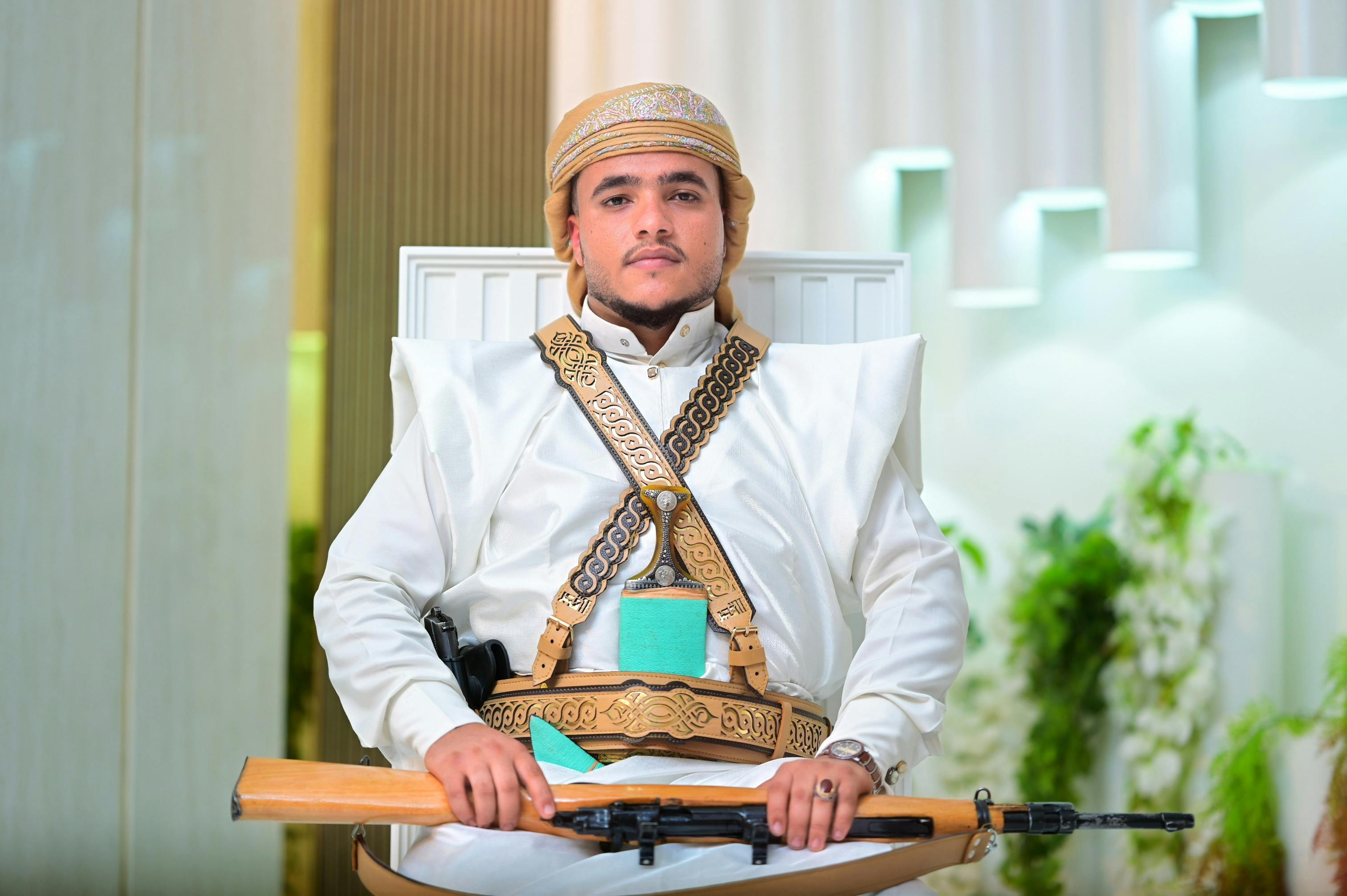 A man in traditional Yemeni attire seated with a rifle indoors, showcasing cultural heritage.