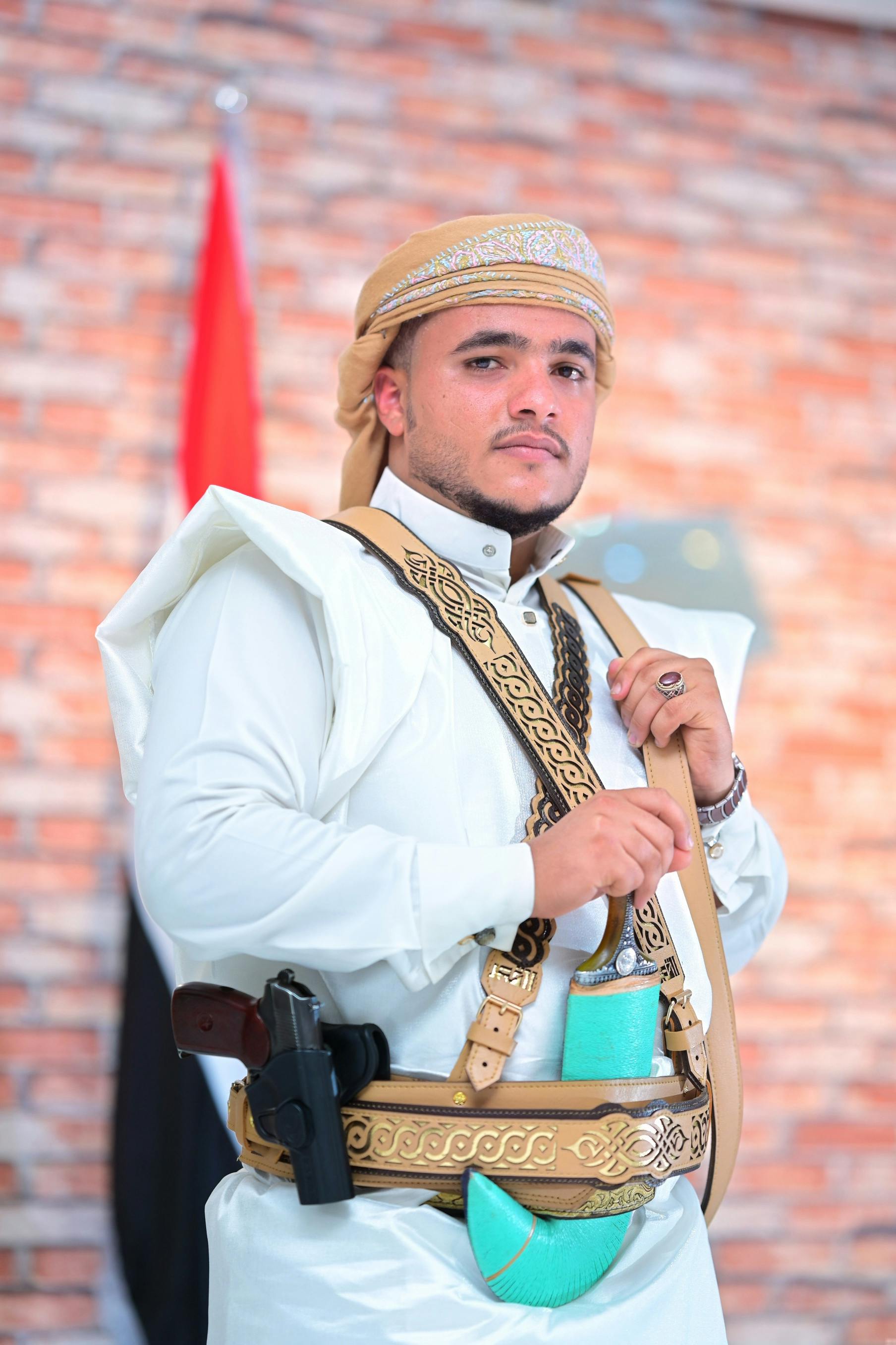 Man in traditional attire with weapon and accessories posing indoors against a brick wall.