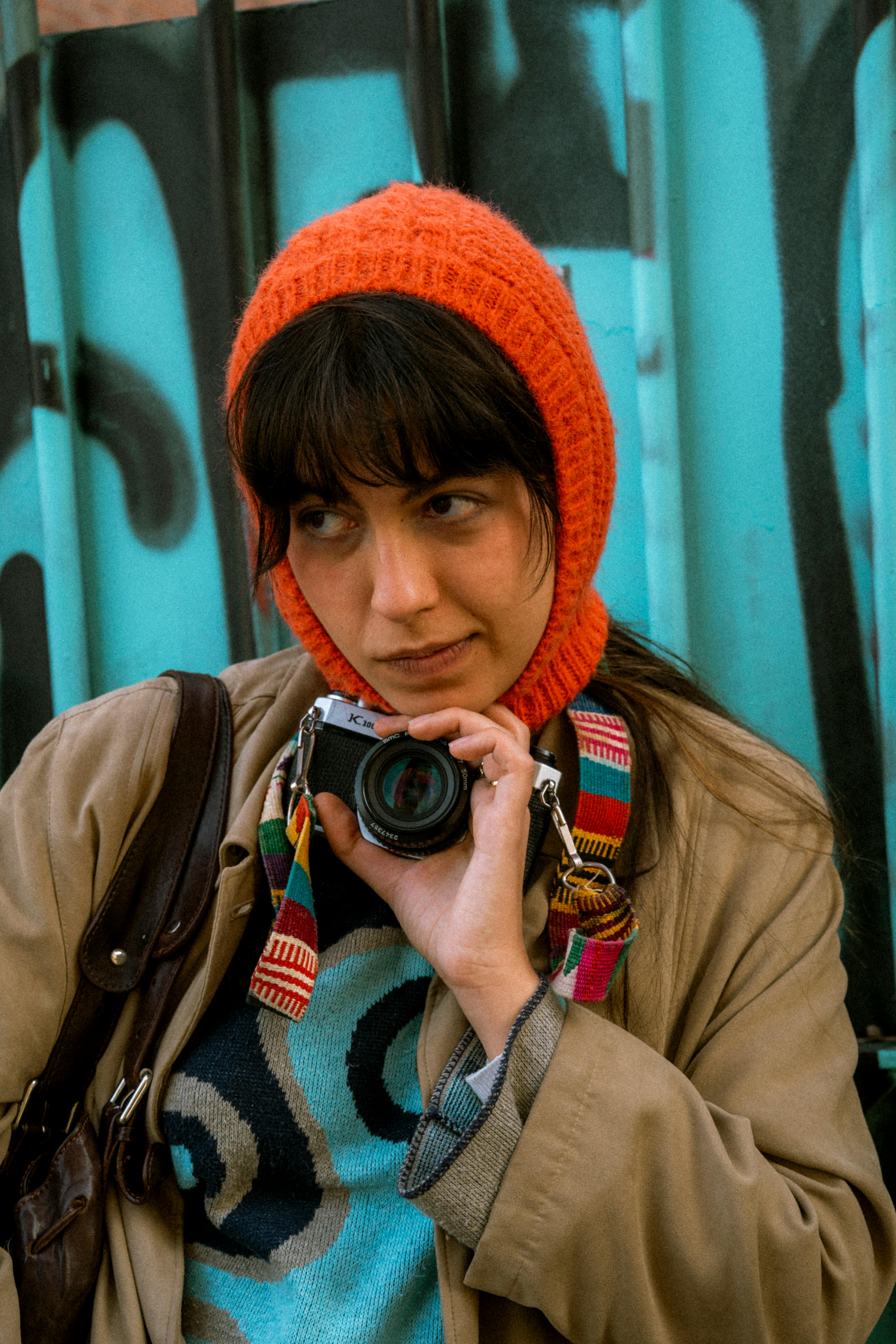 A young woman in warm clothing holding a camera against a graffiti-covered fence.