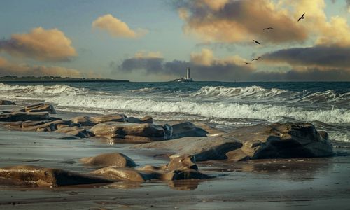 Free Rocky shoreline and seascape with lighthouse and dramatic clouds at sunset. Stock Photo