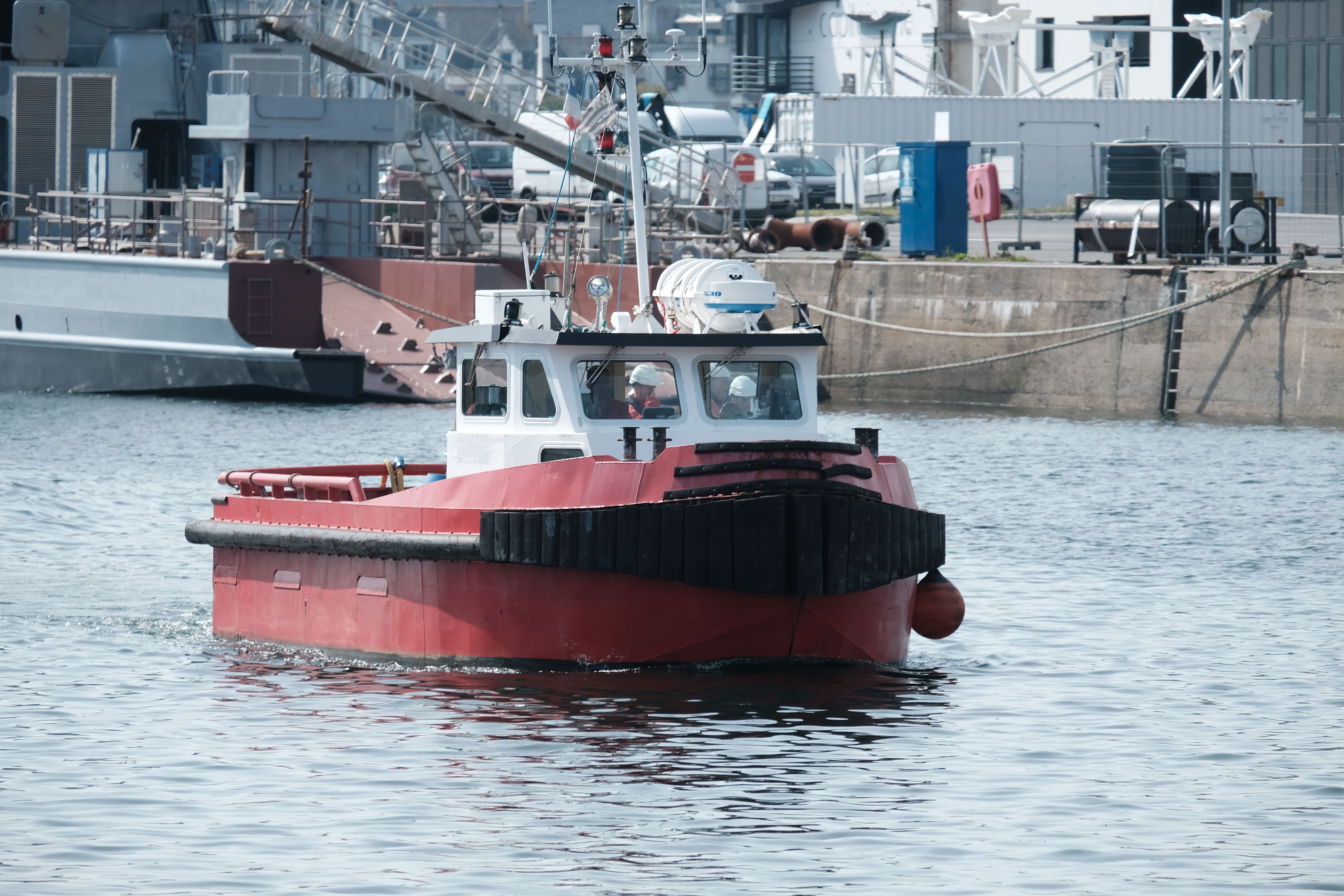 A red tugboat steers through a bustling harbor scene with industrial backdrop.