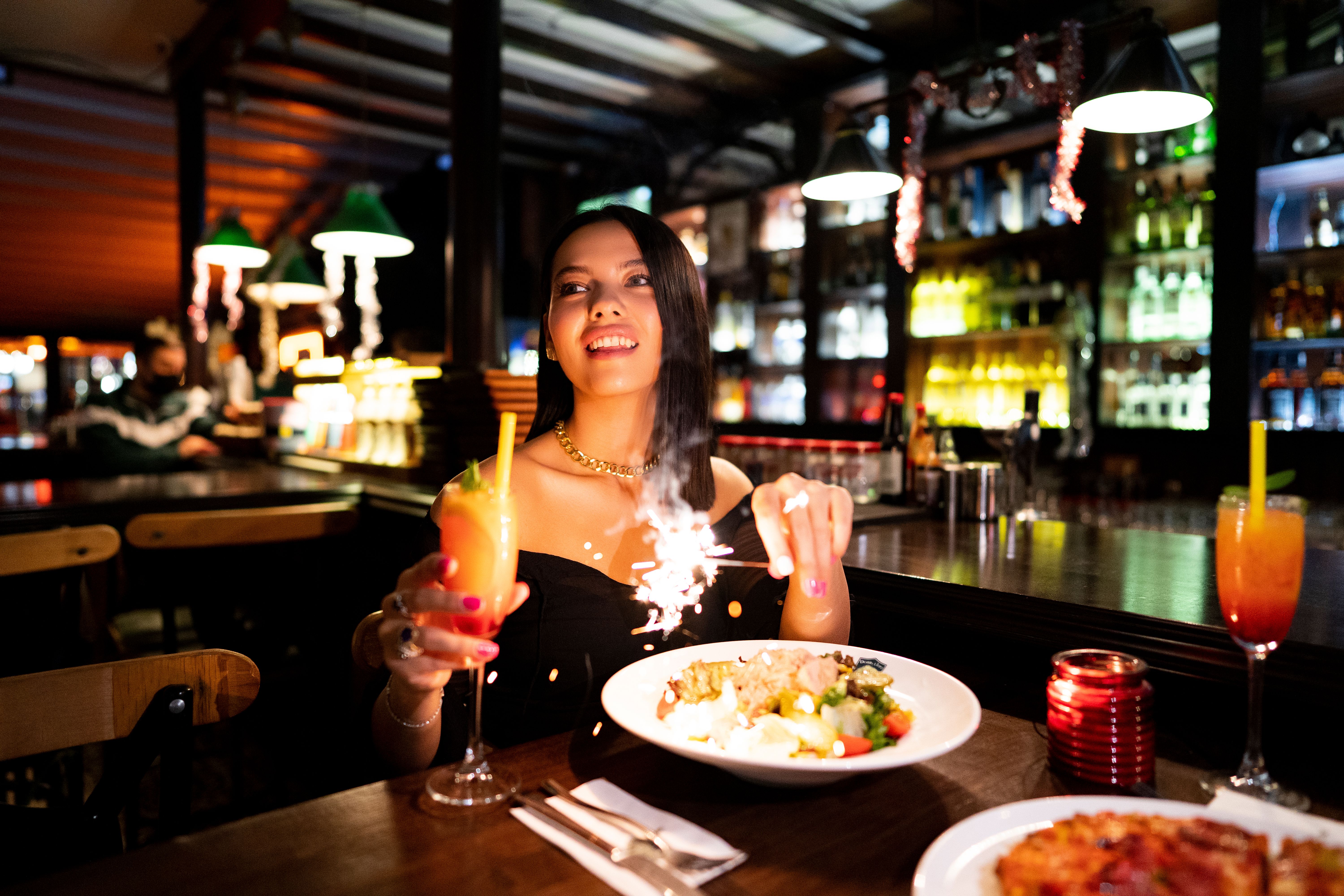 Mulher celebrando com pratos e drinks em um ambiente de bar ou restaurante descontraído, com iluminação acolhedora e decoração moderna.