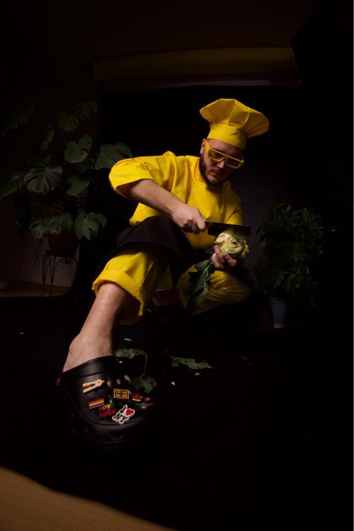 Free A fun, modern portrait of a chef in a bright yellow outfit preparing vegetables on a dark background. Stock Photo
