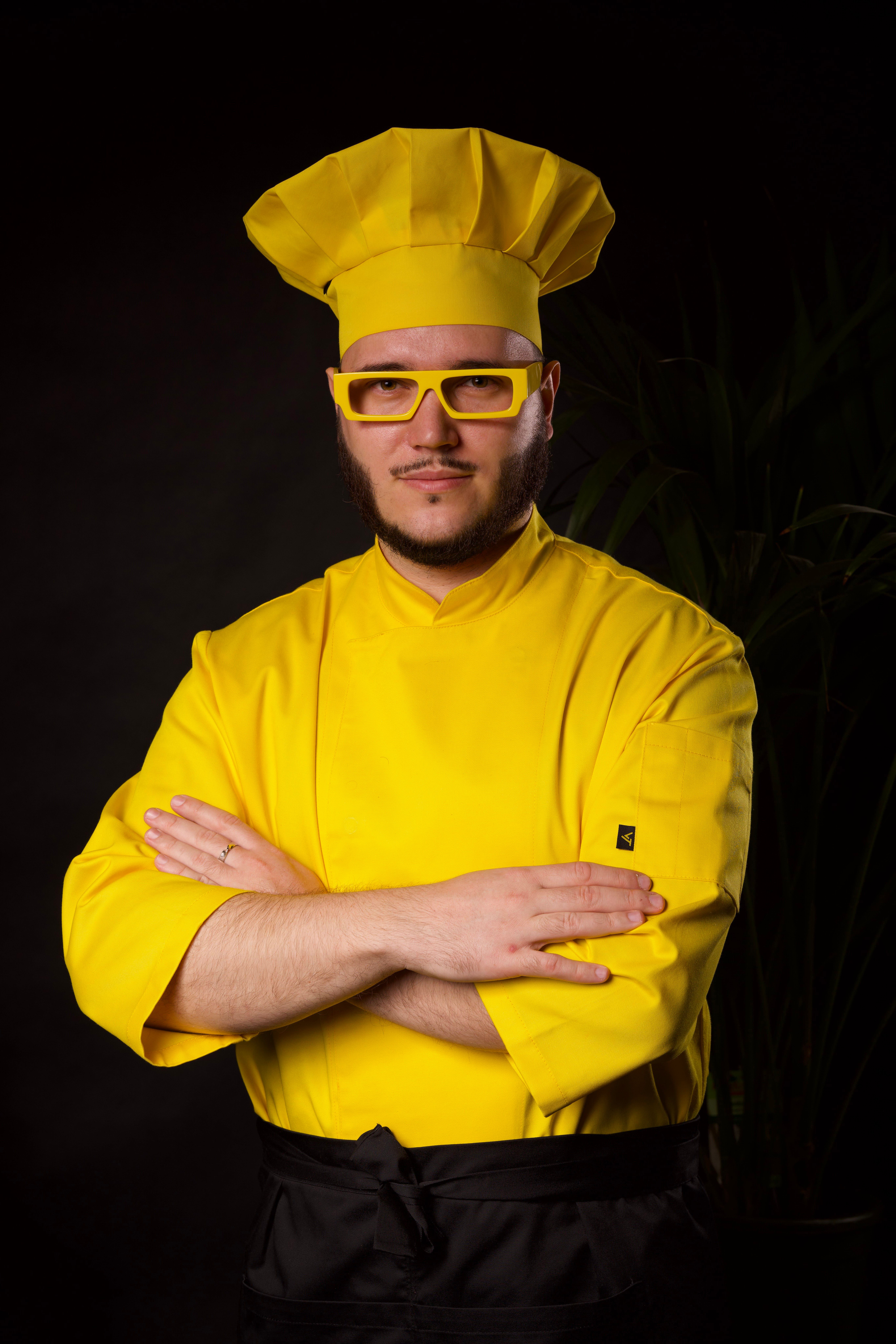Portrait of a professional chef confidently posing in vibrant yellow attire on a black background.