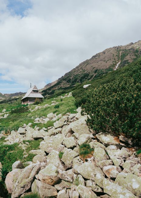 Charming cabin nestled in a rocky, mountainous terrain with lush greenery under a cloudy sky.