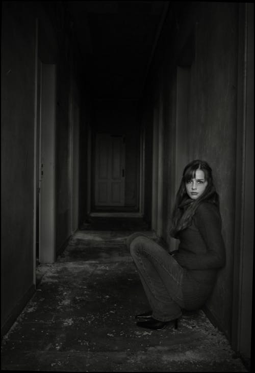 Grayscale Photo of Woman Sitting in Empty Hallway Leaning on Wall
