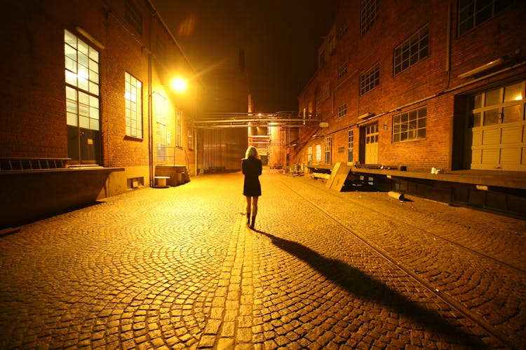 Back View Photo Of Woman Walking Alone In The Middle Of A Cobblestone Alley At Night