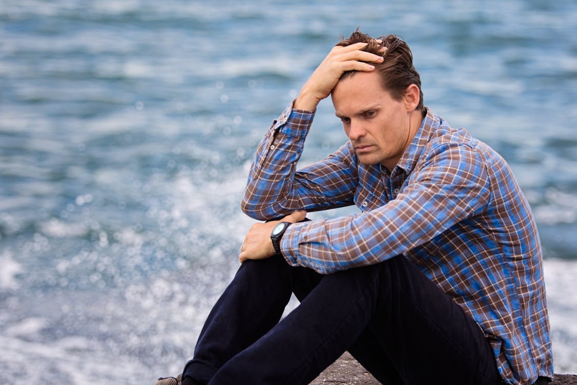 Free Photo of a man sitting near a body of water holding his head Stock Photo