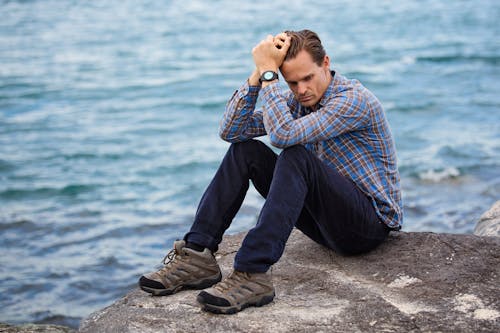 Man wearing blue and maroon plaid shirt sitting on a rock near a body of water