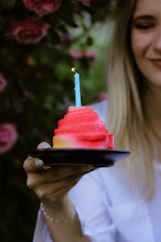 Woman holding a vibrant cupcake with a candle, surrounded by pink flowers.