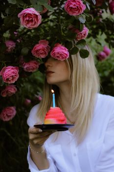 A woman with blonde hair holds a birthday cake with a candle near vibrant pink roses.
