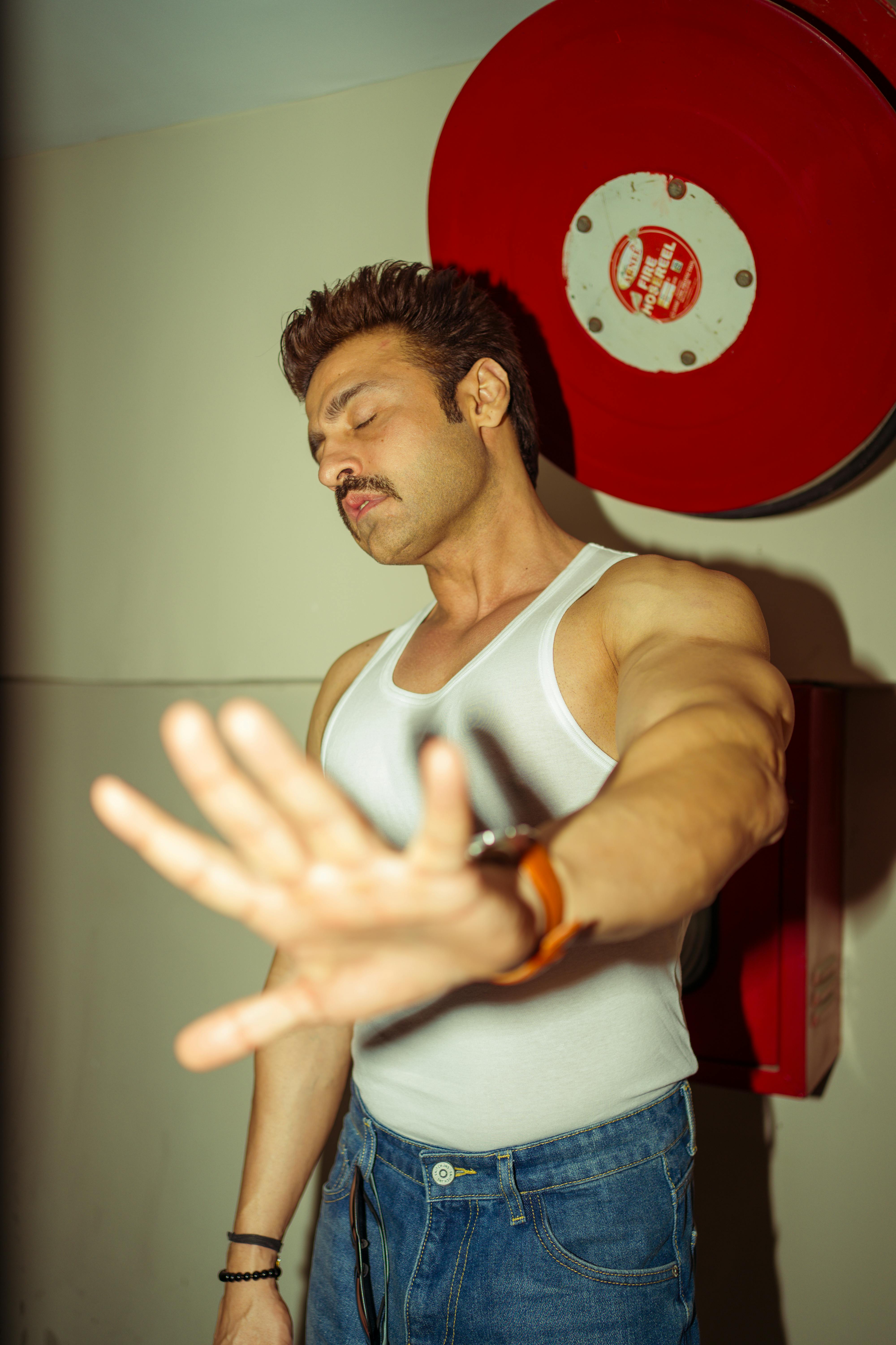 Muscular young man with mustache posing indoors wearing a white tank top and jeans. Stylish and athletic vibe.