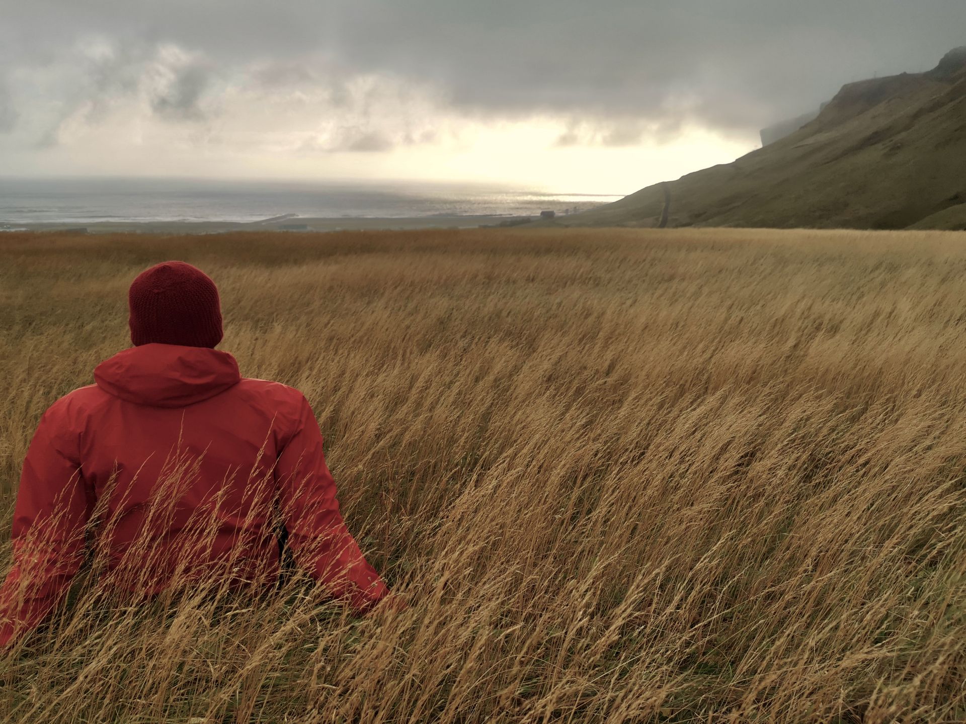 Person in red jacket sits in grassy field near Vík, Iceland, under dramatic sunset skies.