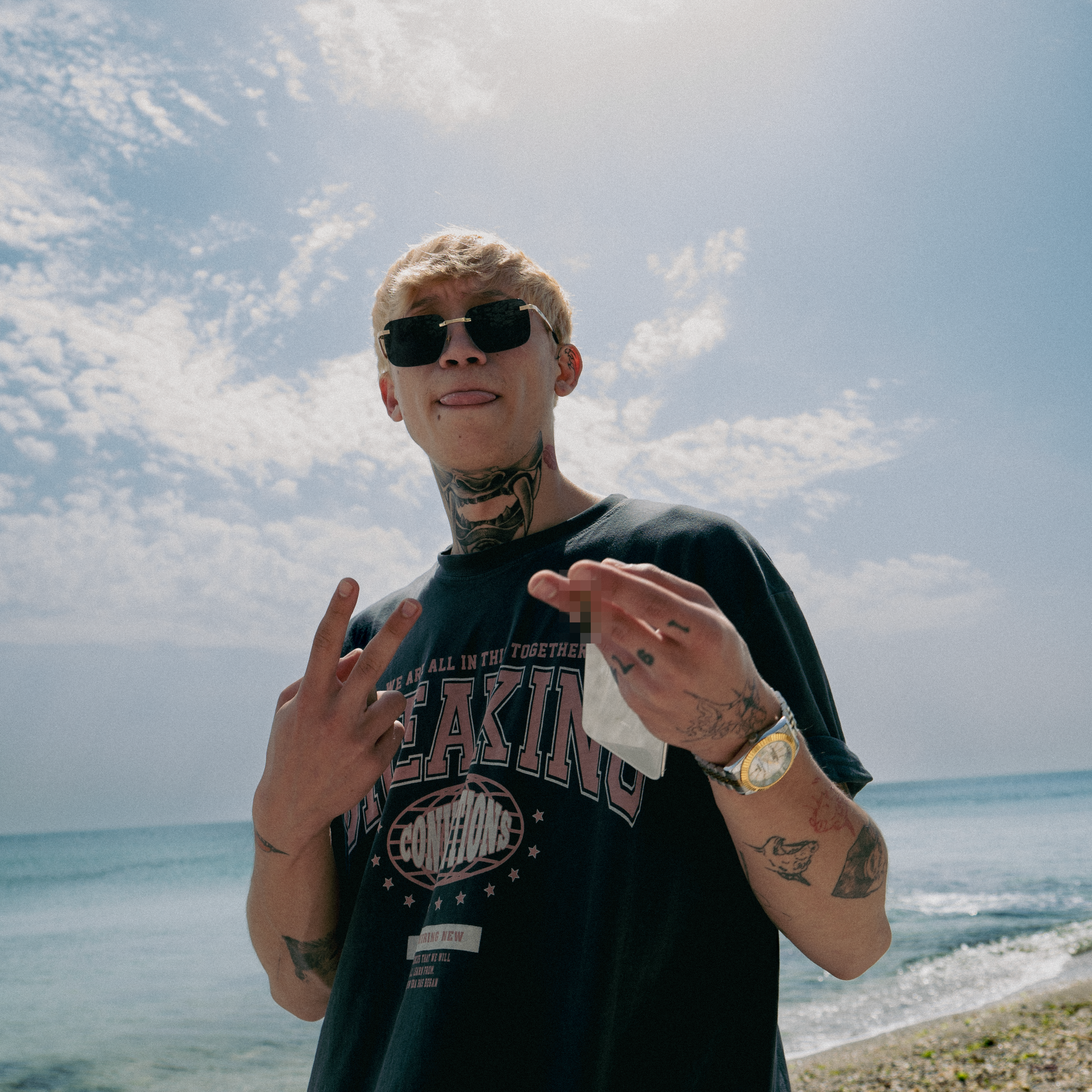Stylish young man with tattoos and sunglasses posing with gesture on a beach under a bright sky.