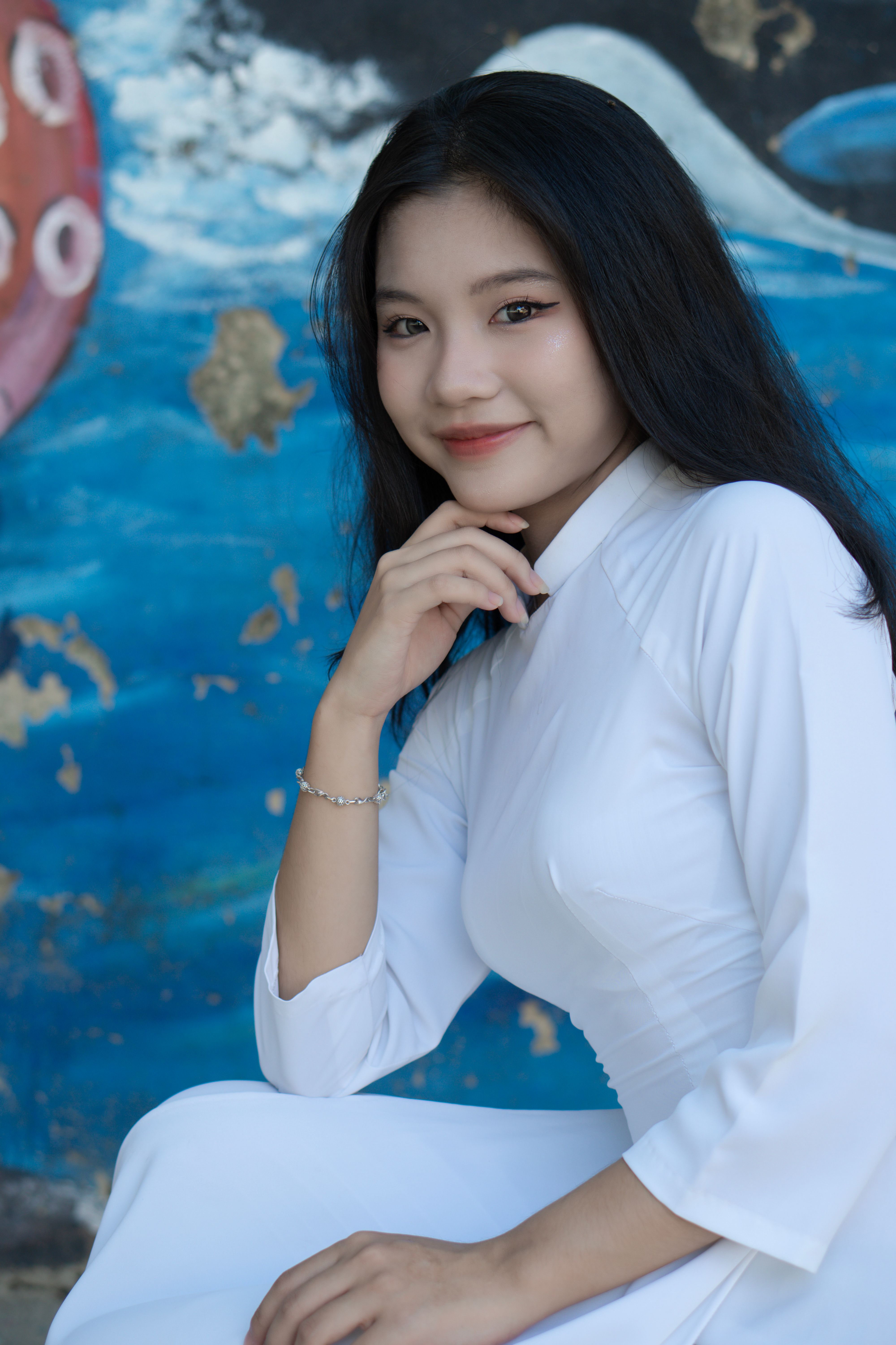 Smiling Asian woman in a white tunic poses in front of a colorful mural, exuding elegance and warmth.