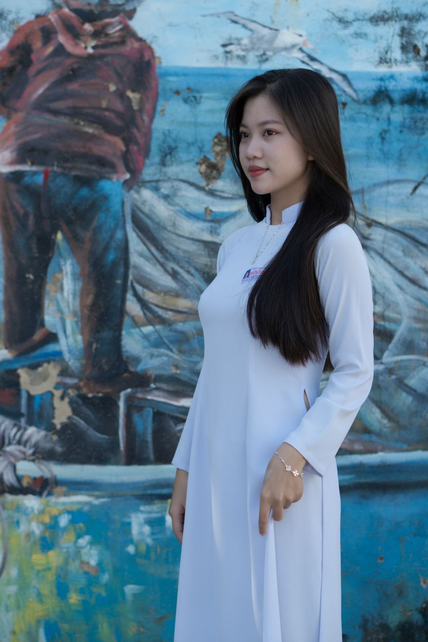 Portrait of an Asian woman standing against a colorful mural, wearing a traditional white dress.