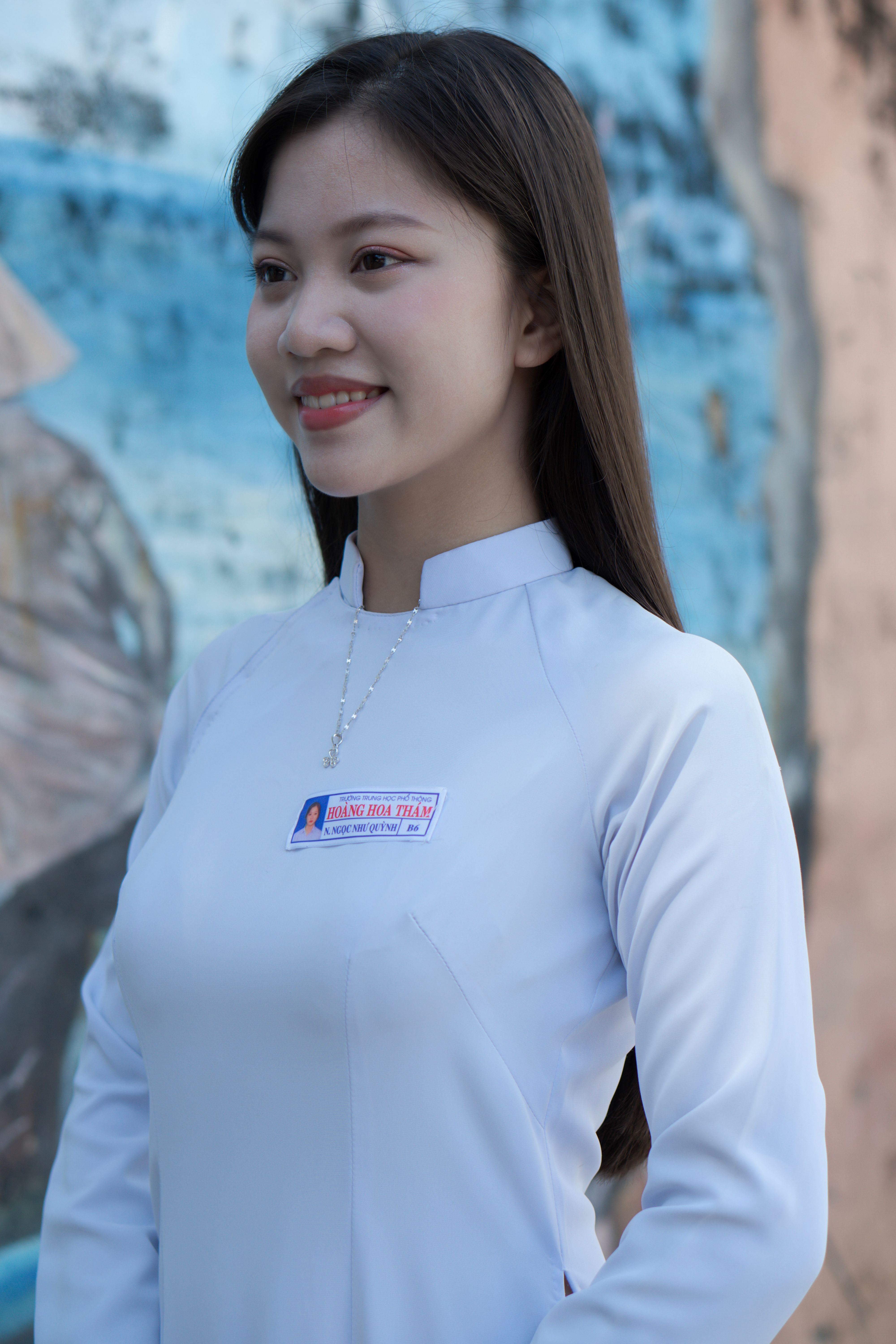A young woman with long hair smiles softly in a white traditional outfit, outdoors with a blurred background.