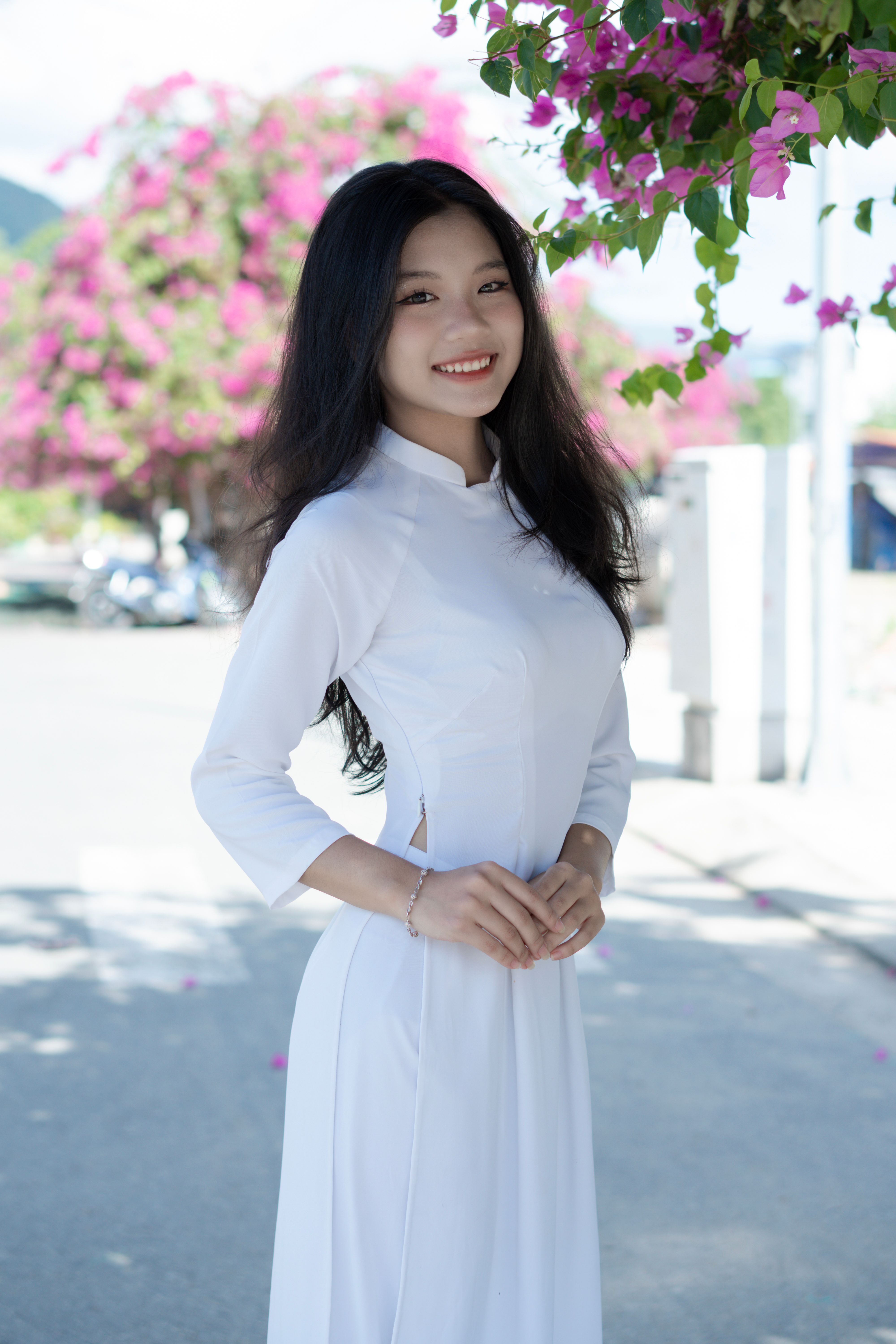 Smiling woman in white ao dai standing outdoors with vibrant flowers.