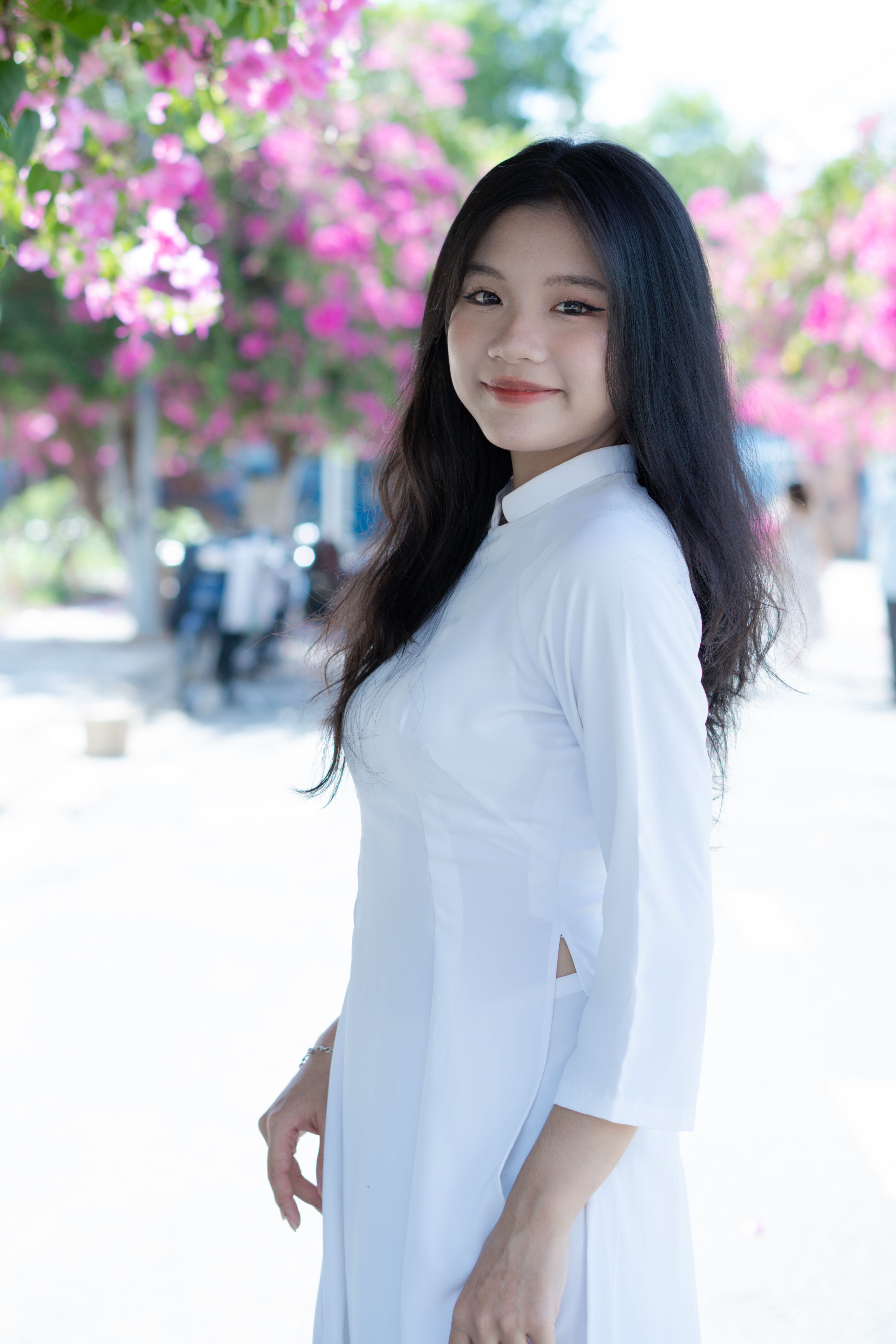Beautiful portrait of an Asian woman in a white dress, standing confidently outdoors with pink flowe