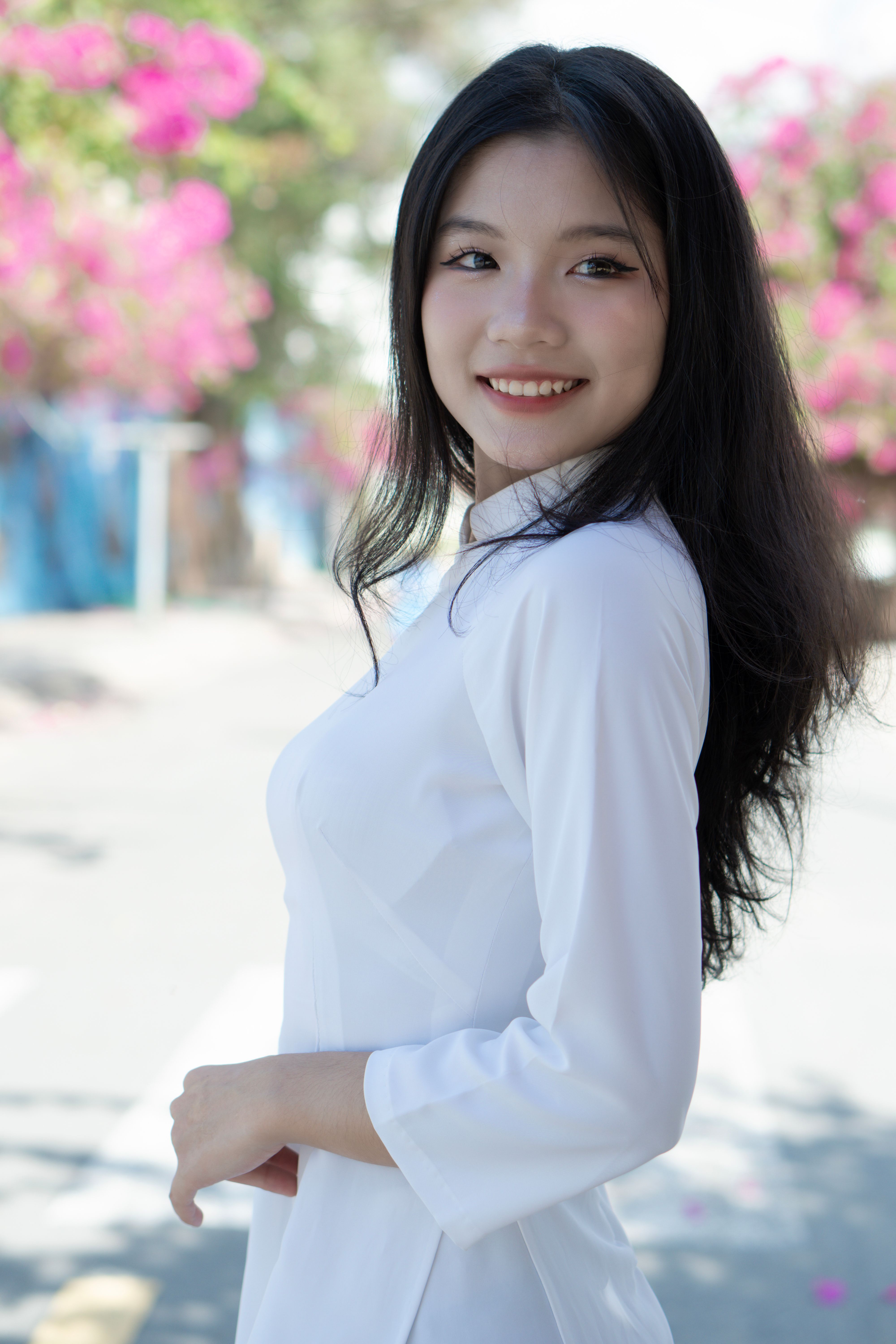 Portrait of a smiling Asian woman in a traditional white dress, standing outdoors with pink flowers in the background.