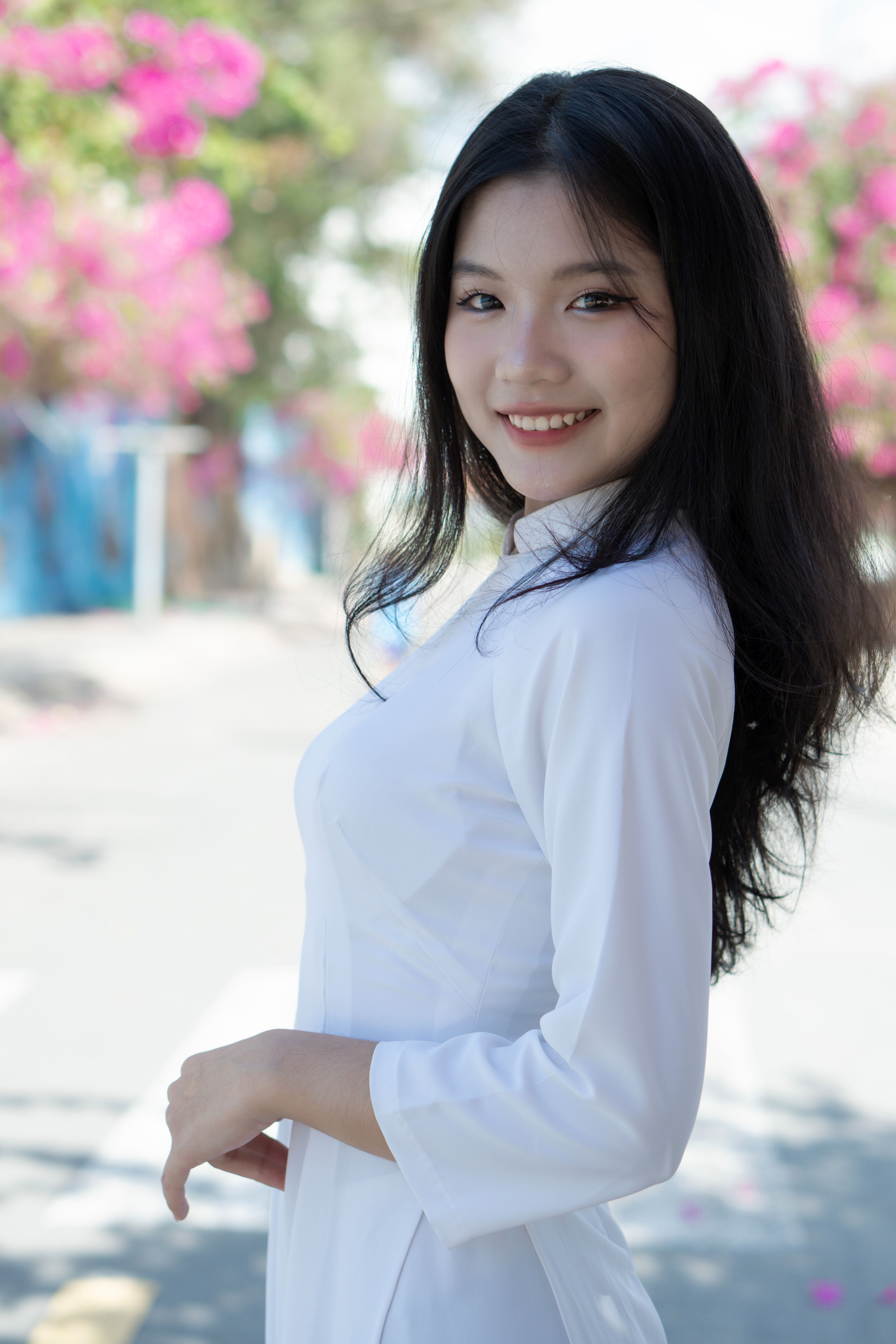 A smiling woman in traditional white dress poses outdoors with pink flowers in the background.
