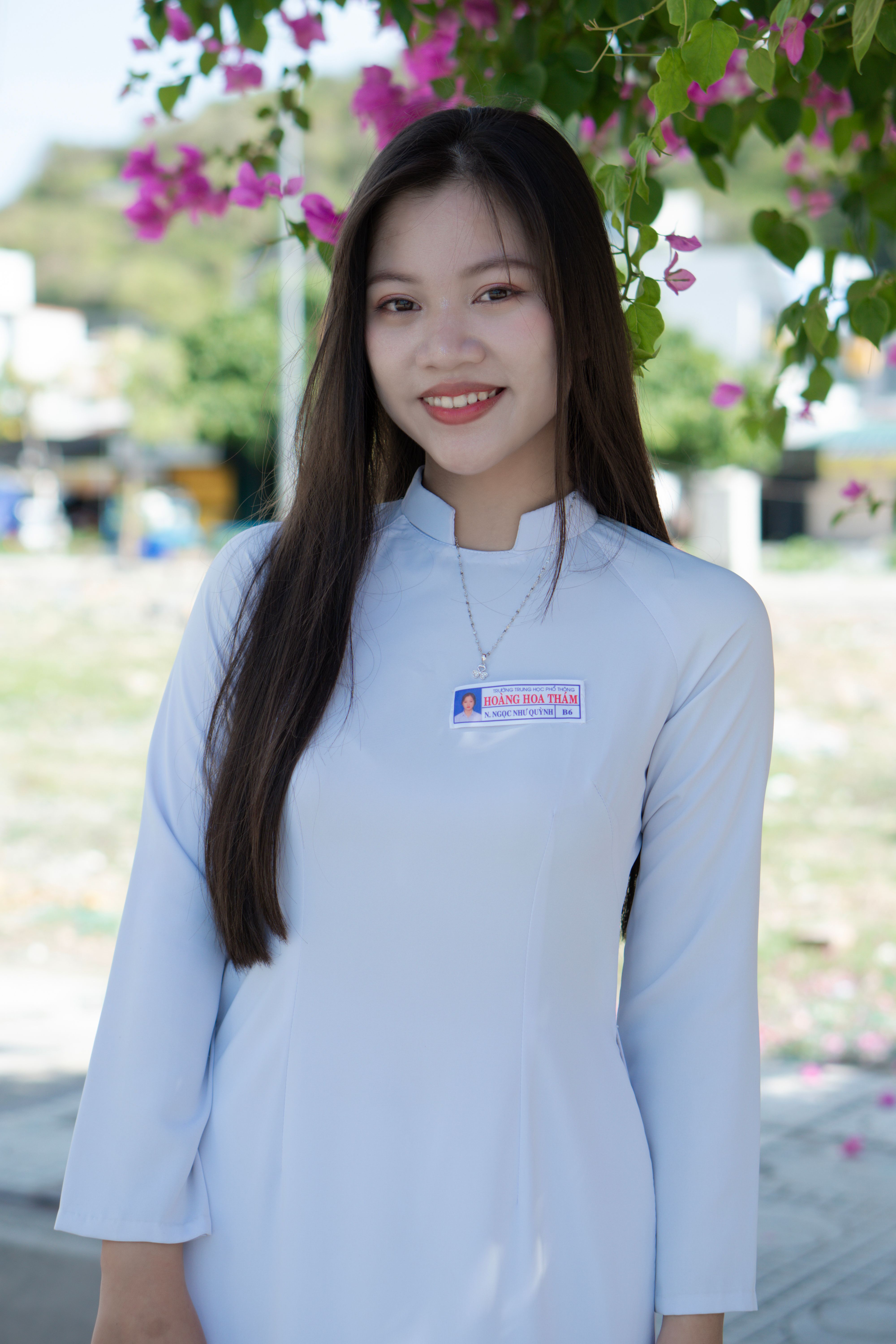 Portrait of an Asian woman smiling in traditional white dress outdoors under blooming flowers.