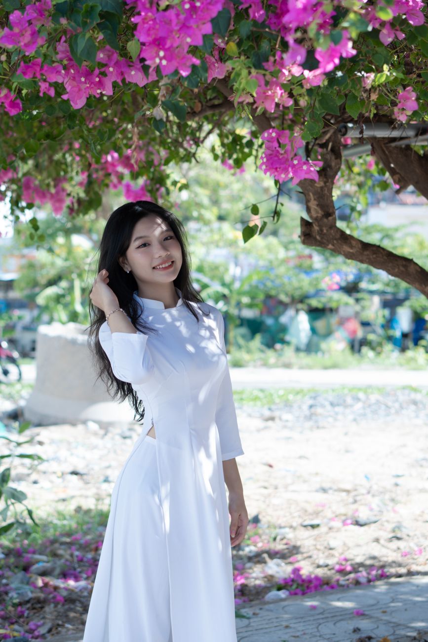 Asian woman in traditional white dress under pink blossoms, smiling outdoors.