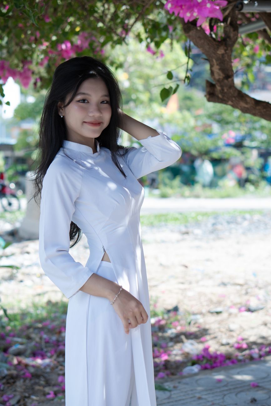 Asian woman in a white ao dai standing gracefully under a tree with vibrant pink blossoms, exuding elegance and charm.