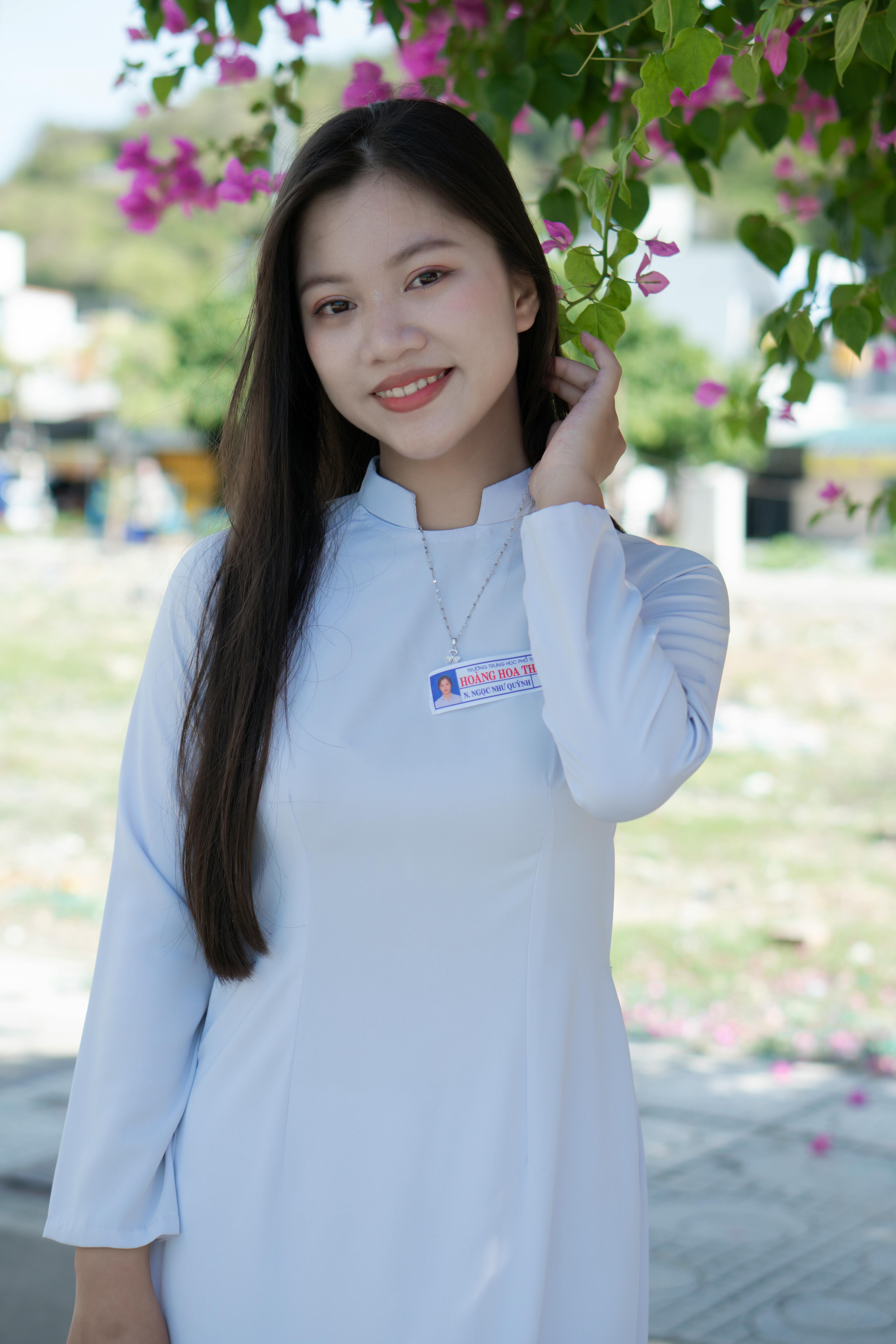 A smiling woman in traditional clothing poses outdoors with blooming flowers.