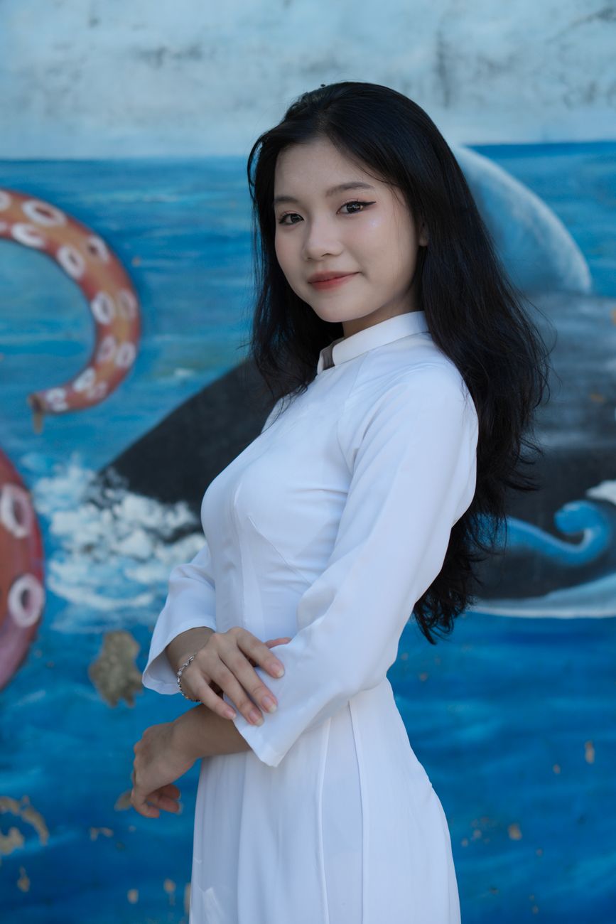 Portrait of a woman in a white ao dai, standing confidently against a colorful mural background.